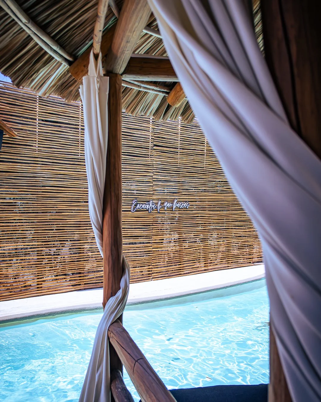 Vista desde una cabaña de madera con cortinas blancas hacia una piscina y un fondo de muro de madera con una frase en español. day pass beach club de playa pie de la cuesta, hotel boutique baXar acapulco