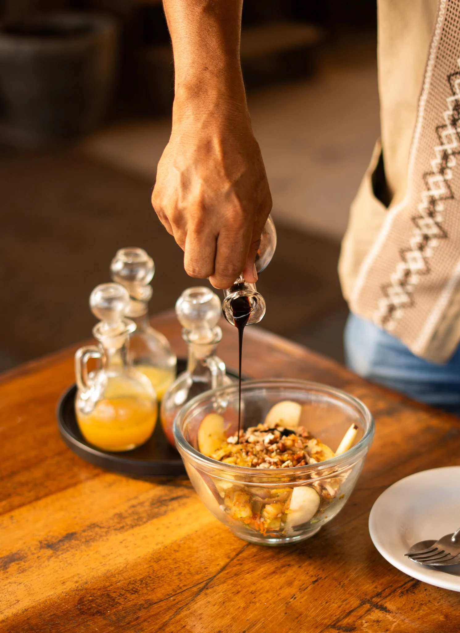 Persona vertiendo vinagre en una ensalada en un cuenco de vidrio, con ingredientes como plátanos y nueces, en una mesa de madera. Restaurante Xepina, hotel baXar playa Pie de la cuesta Acapulco.