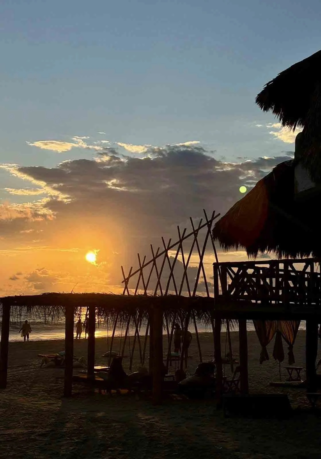 Atardecer en la playa con estructura de madera y techo de palma, personas caminando y sentadas, cielo con nubes y sol en el horizonte