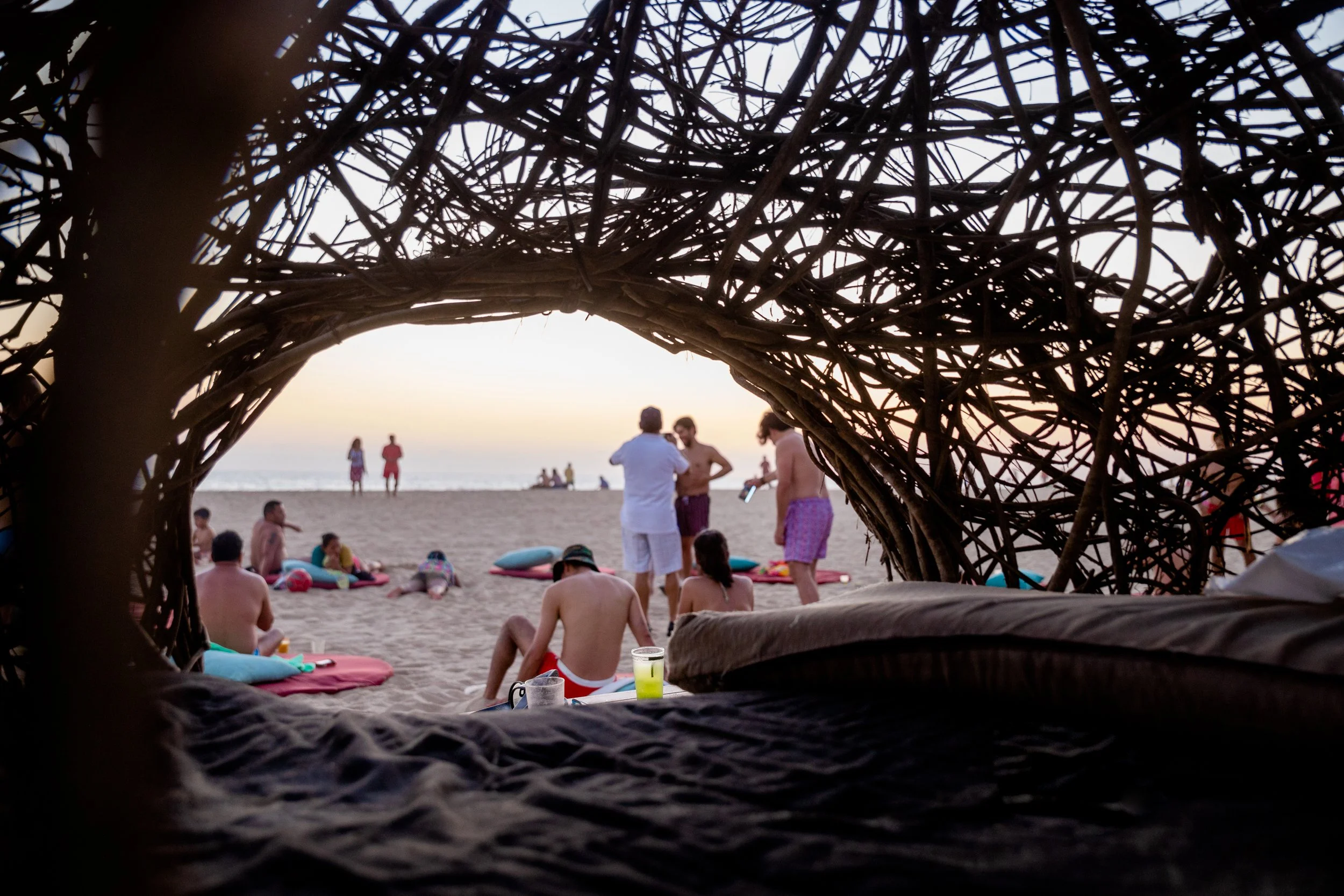 Vista desde dentro de una estructura de ramas de árbol o palapa en una playa, mirando hacia una multitud de personas de espaldas, algunas en la arena y otras en el agua, disfrutando del atardecer en el mar club de playa pie de la cuesta, hotel baXar