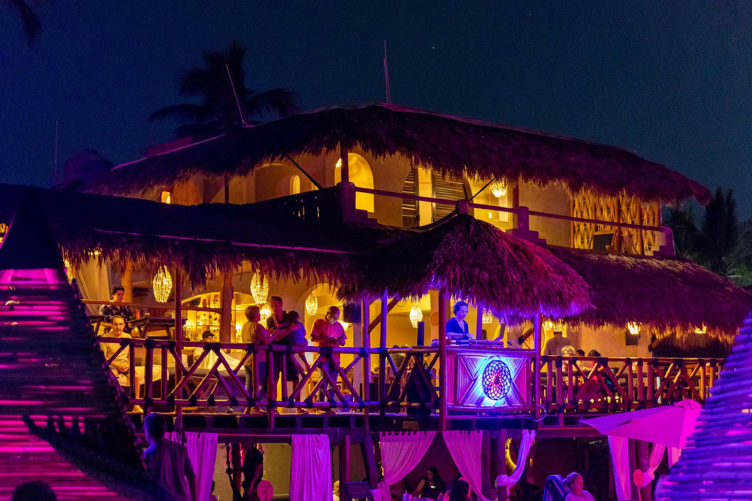 Escena nocturna en un rrestaurante club de playa pie de la cuesta, bar hotel baXar con decoraciones de estilo tropical, con palmeras y techos de palma, luces de colores y personas disfrutando en un ambiente alegre y relajado.