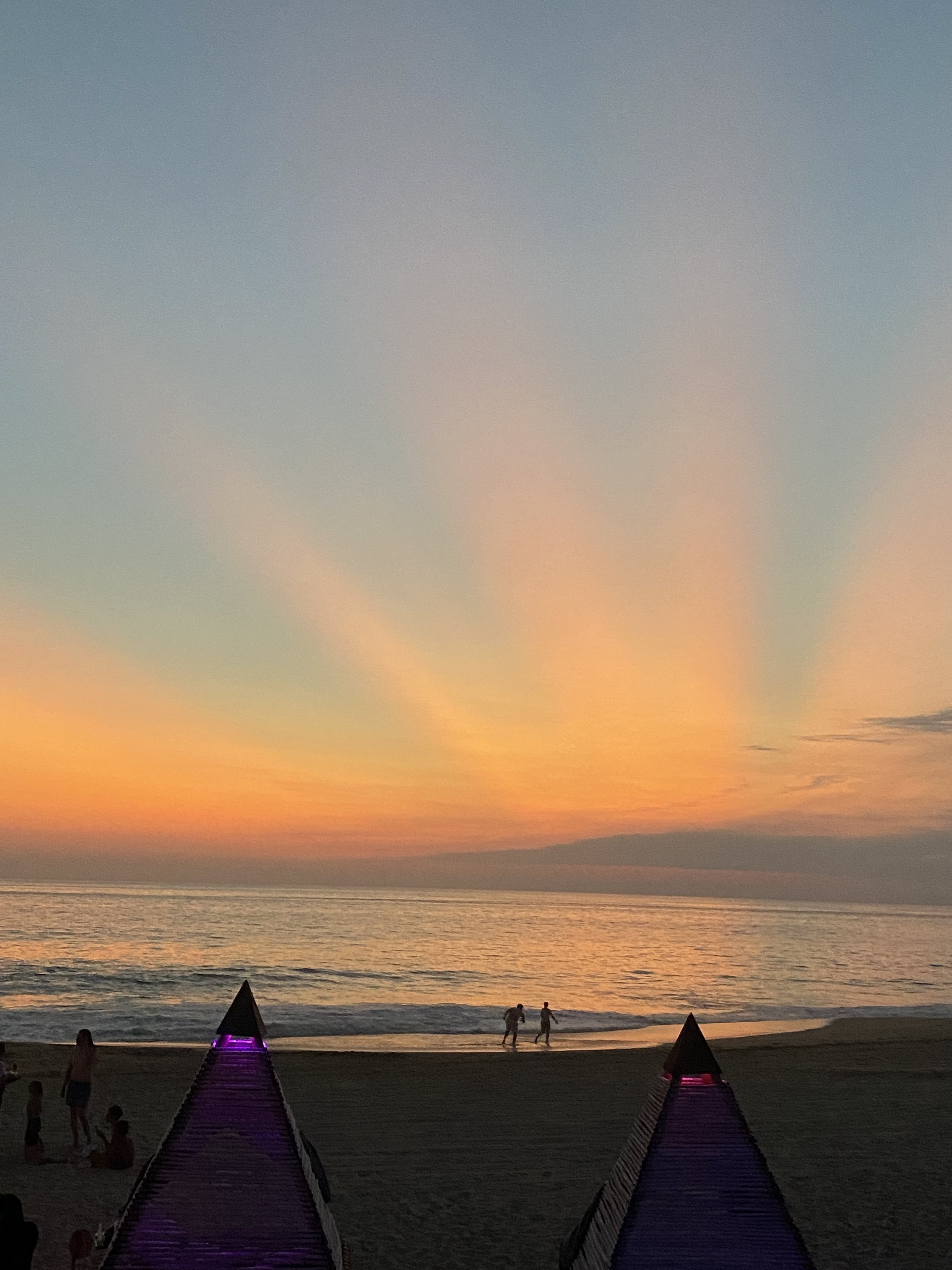 Atardecer en la playa con sombrillas en primer plano, dos personas caminando por la orilla del beach club del hotel baXar club de playa pie de la cuesta, atardecer un cielo con tonos rosados, naranjas y azules. 