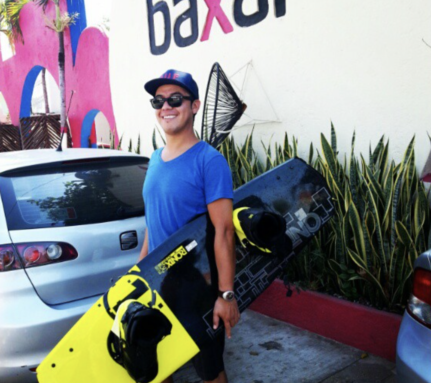 DJ Shine sonriendo con gafas y gorra, sosteniendo una tabla de snowboard de color negro y amarillo, en un estacionamiento junto a un coche y plantas ornamentales, con un mural colorido en la pared de fondo. Hotel baXar pie de la cuesta