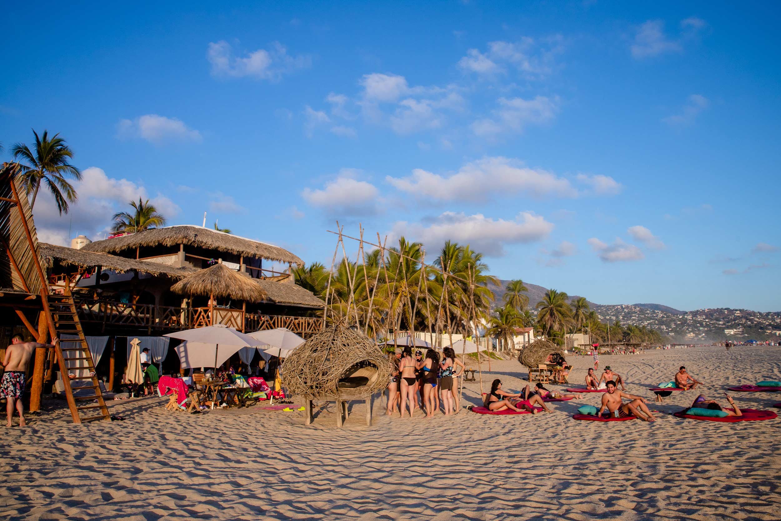 Playa con palmeras, sombrillas y gente disfrutando del sol, con estructuras de madera con techos de paja y un cielo despejado con algunas nubes. beach club del hotel baXar club de playa pie de la cuesta