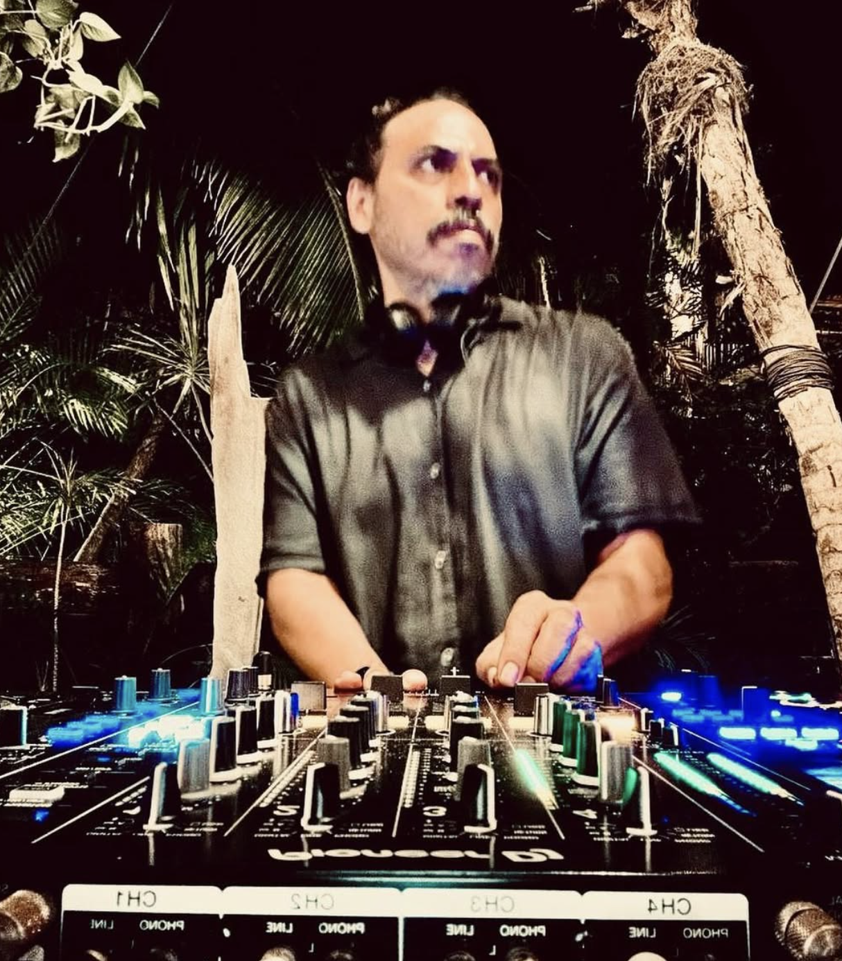 Un DJ tocando en una cabina de mezclas en un entorno con plantas y palmeras, durante la noche.