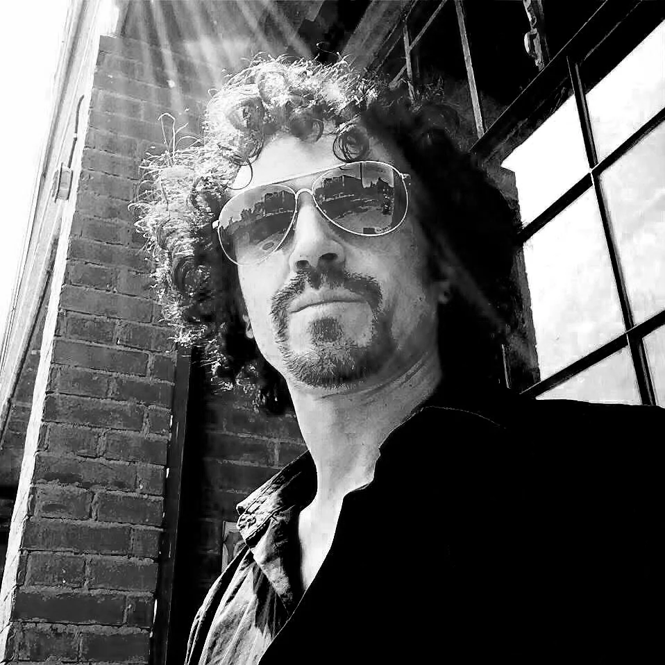 Retrato en black y white de un DJ JABA Joao Adilson Balderas - cabello rizado y barba, usando gafas de sol que reflejan edificios y cielo, en un entorno urbano con una pared de ladrillos y ventanas de metal.