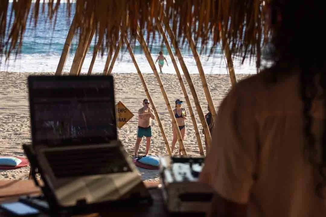 Vista desde dentro de una cabaña en la playa, donde se observa a varias personas bañándose y tomando el sol, con la vista del mar y la arena en el fondo.