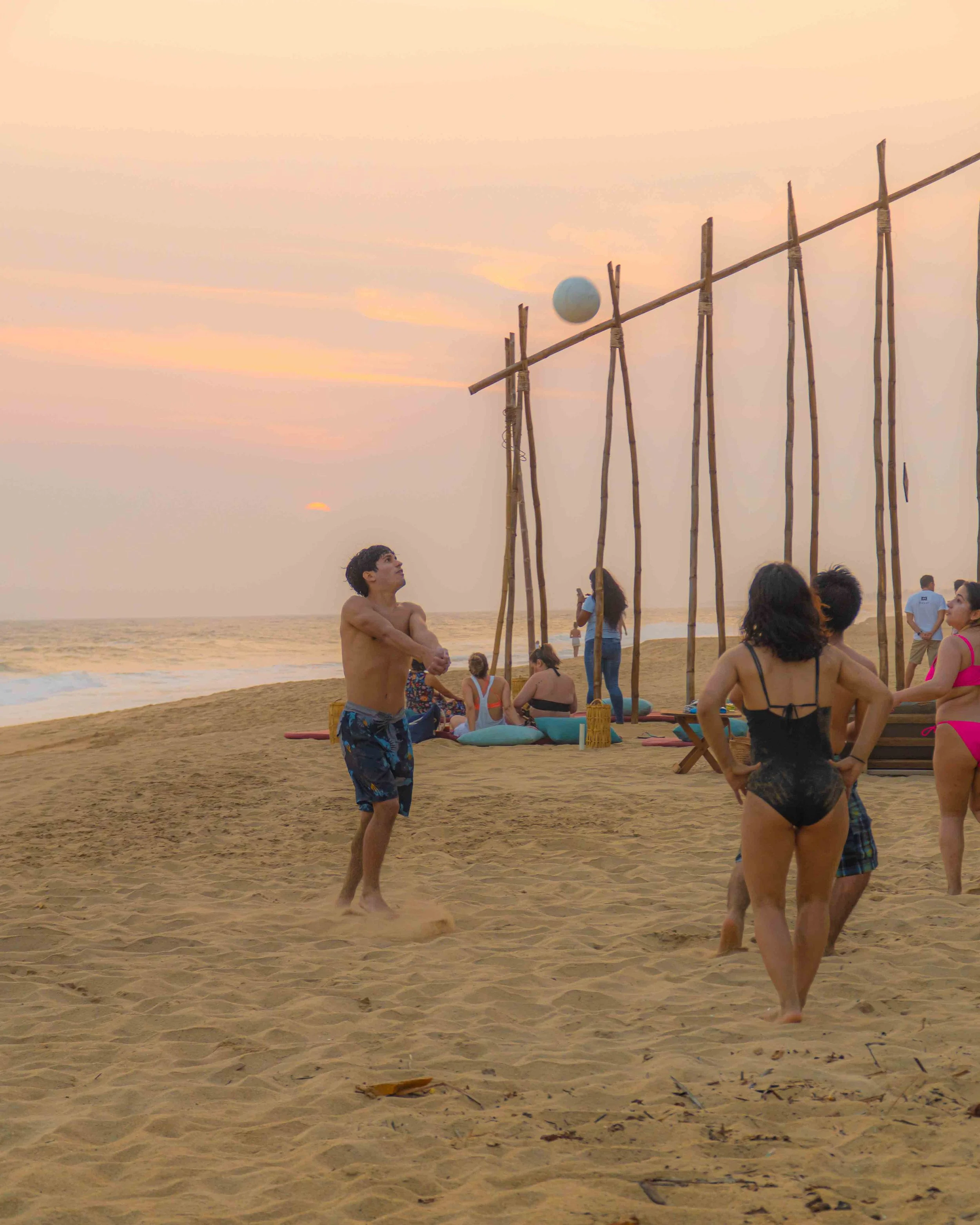 Personas jugando volibol en la playa al atardecer, con estructura de madera en el fondo y varias personas descansando en la arena. hotel baxar pie de la cuesta 