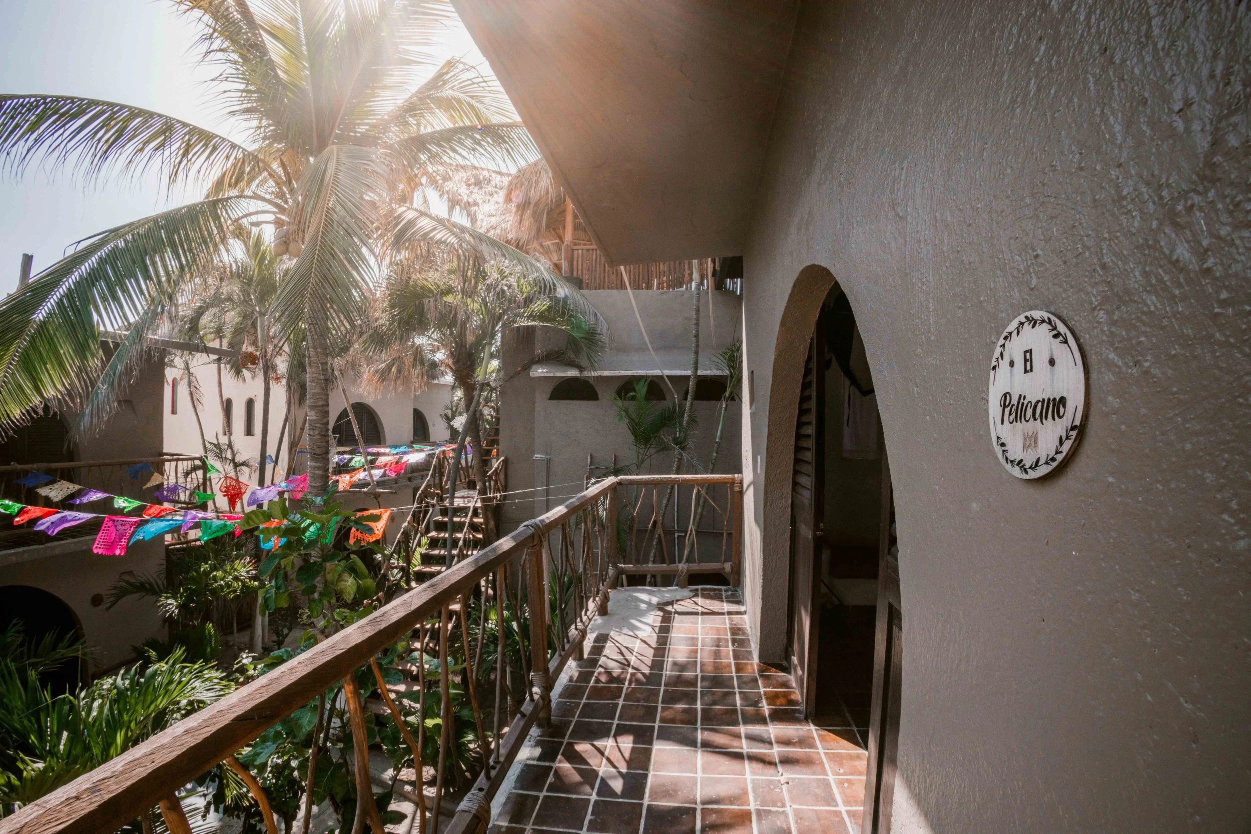 Balconada con piso de baldosas de barro, barandilla de madera, vista a un patio con palmeras y banderines de papel de colores, en un edificio de estilo mexicano con paredes de color beige y letrero que dice ‘El Pelícano’ en la pared.