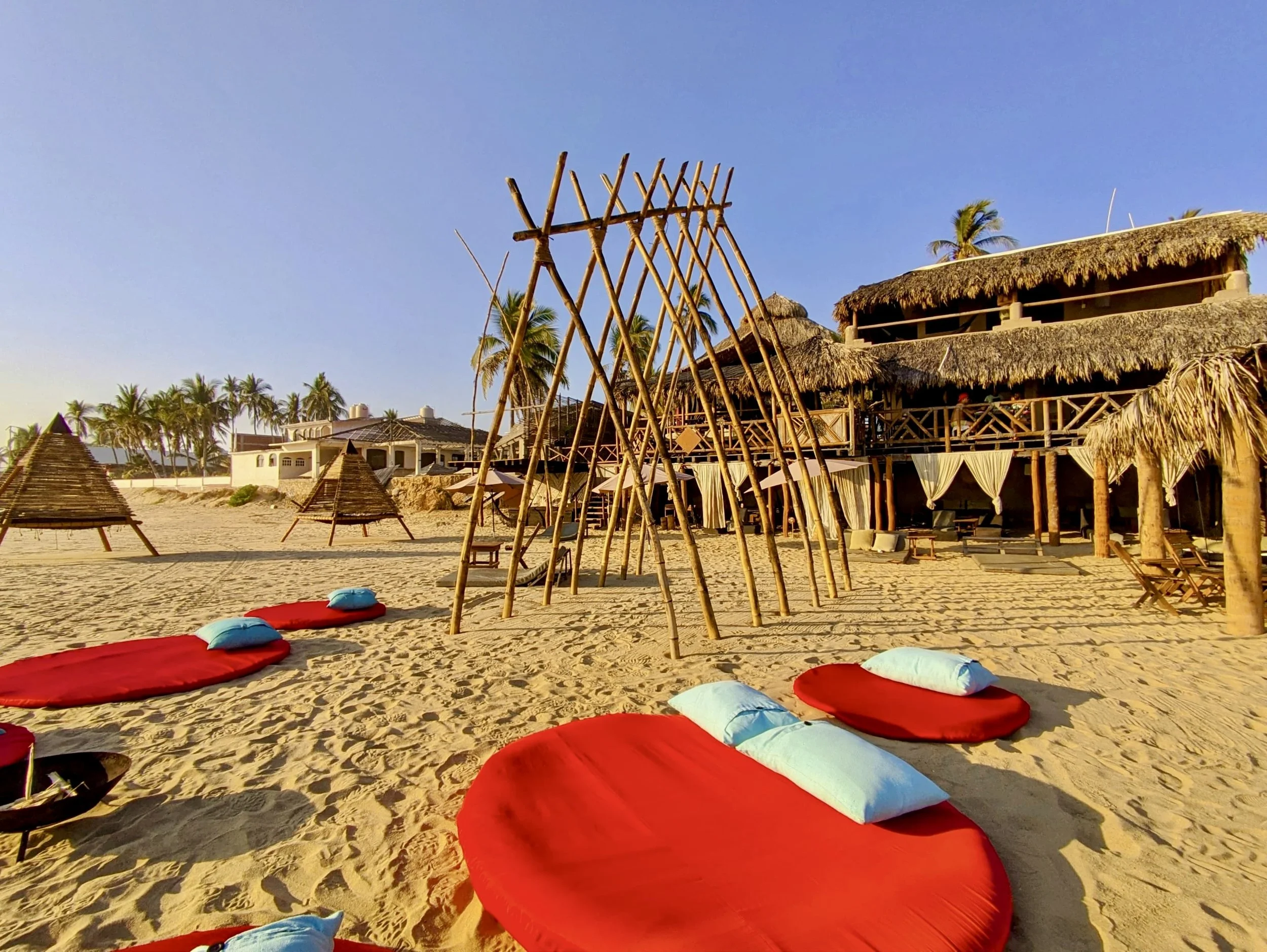 Playas de arena con colchones y cojines rojos y blancos, estructuras de madera con techos de paja, palmeras y un edificio con balcón y techo de paja, cielo despejado y soleado. Playa Pie de la Cuesta, Acapulco, Hotel baXar