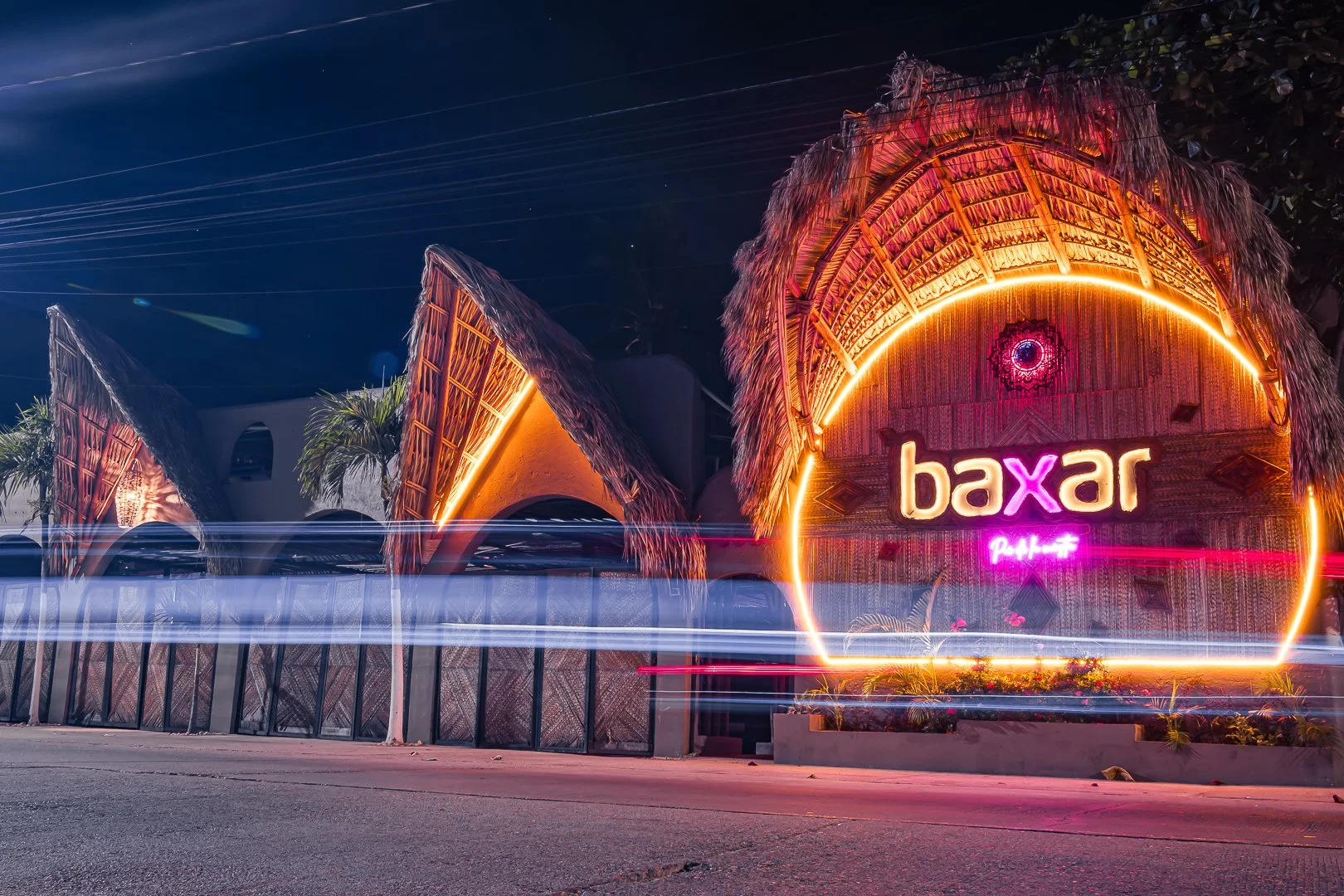 Entrada del hotel Baxar, con cartel luminoso en letras neón rosas y anaranjadas y construcciones de techos de paja iluminados con luces de colores, noche con cielo oscuro y tráfico en movimiento. boutique playa pie de la cuesta, baXar Acapulco