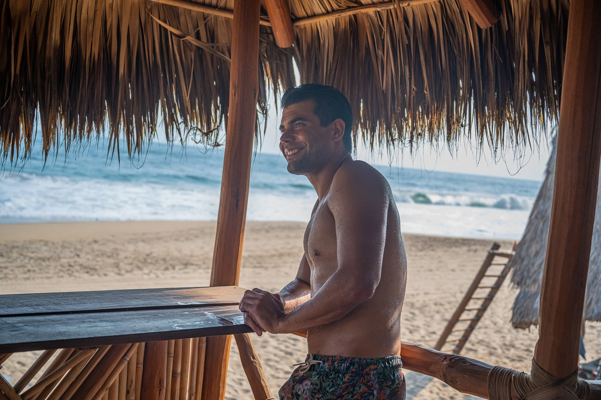 Un hombre en la playa, bajo un techo de palapa, sonriente, con el mar de fondo.  club de playa pie de la cuesta, hotel boutique baXar acapulco