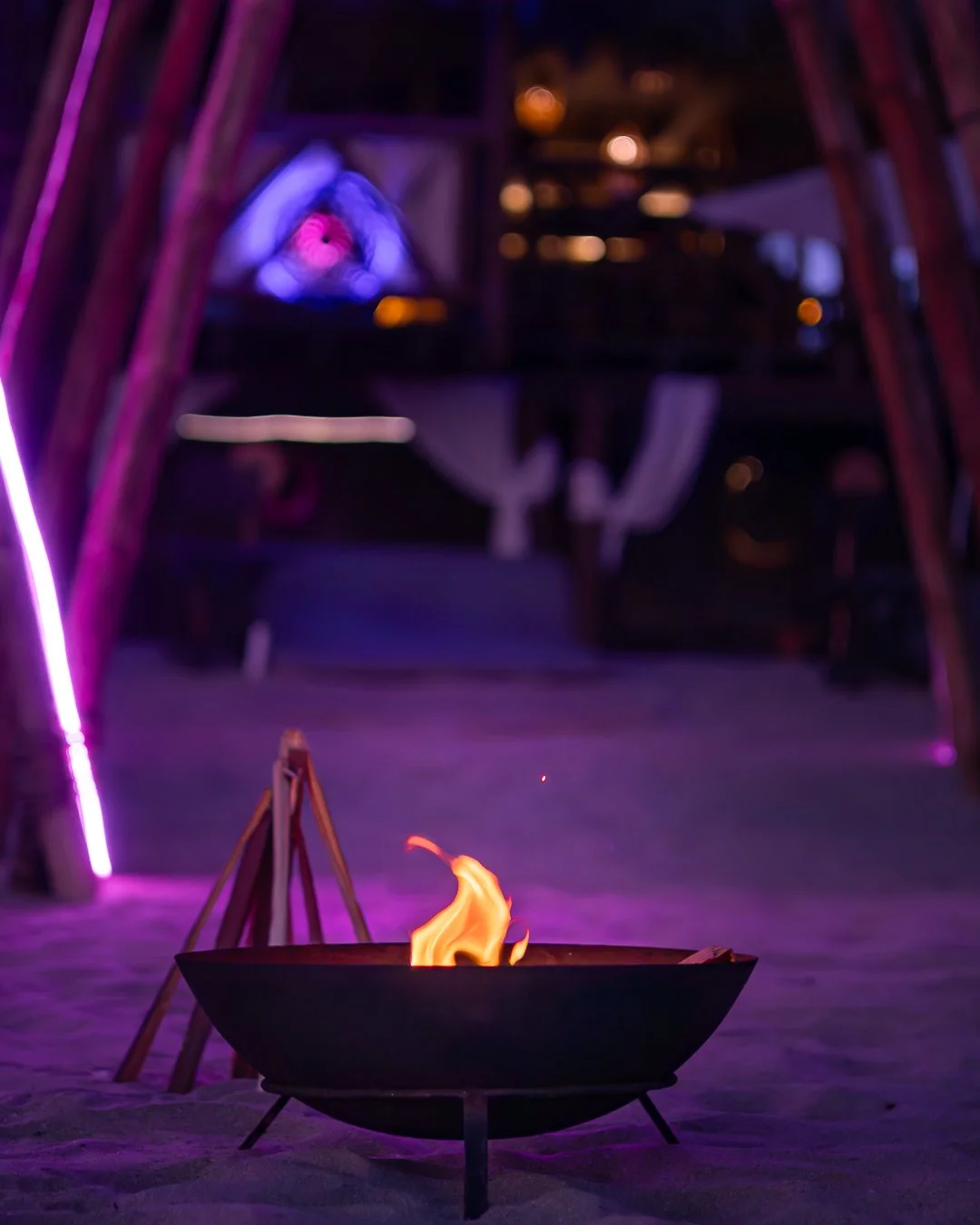 Fuego en una parrilla de carbón en primer plano, en un ambiente iluminado con luces moradas y rosadas, con una estructura de madera y un área de descanso al fondo.