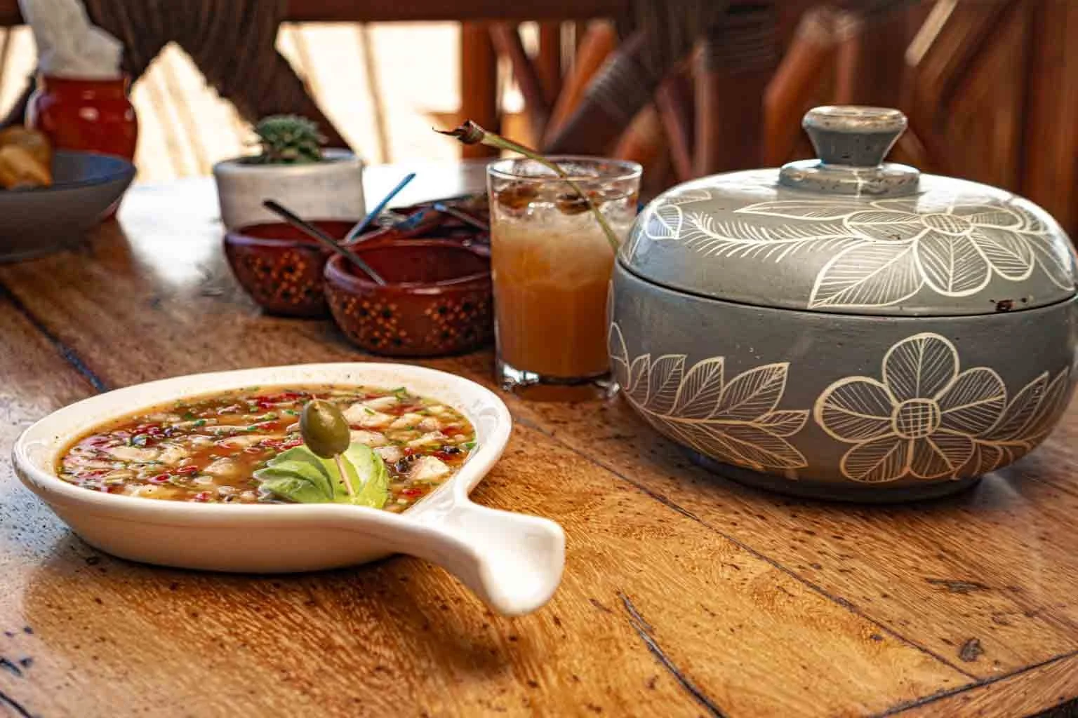 Imagen de una mesa de madera con una sopa en un plato con un aceituna y una hoja de albahaca, un vaso con una bebida fría y decorada con una flor, y un recipiente de cerámica con diseño floral, en un ambiente de interior con luz natural.