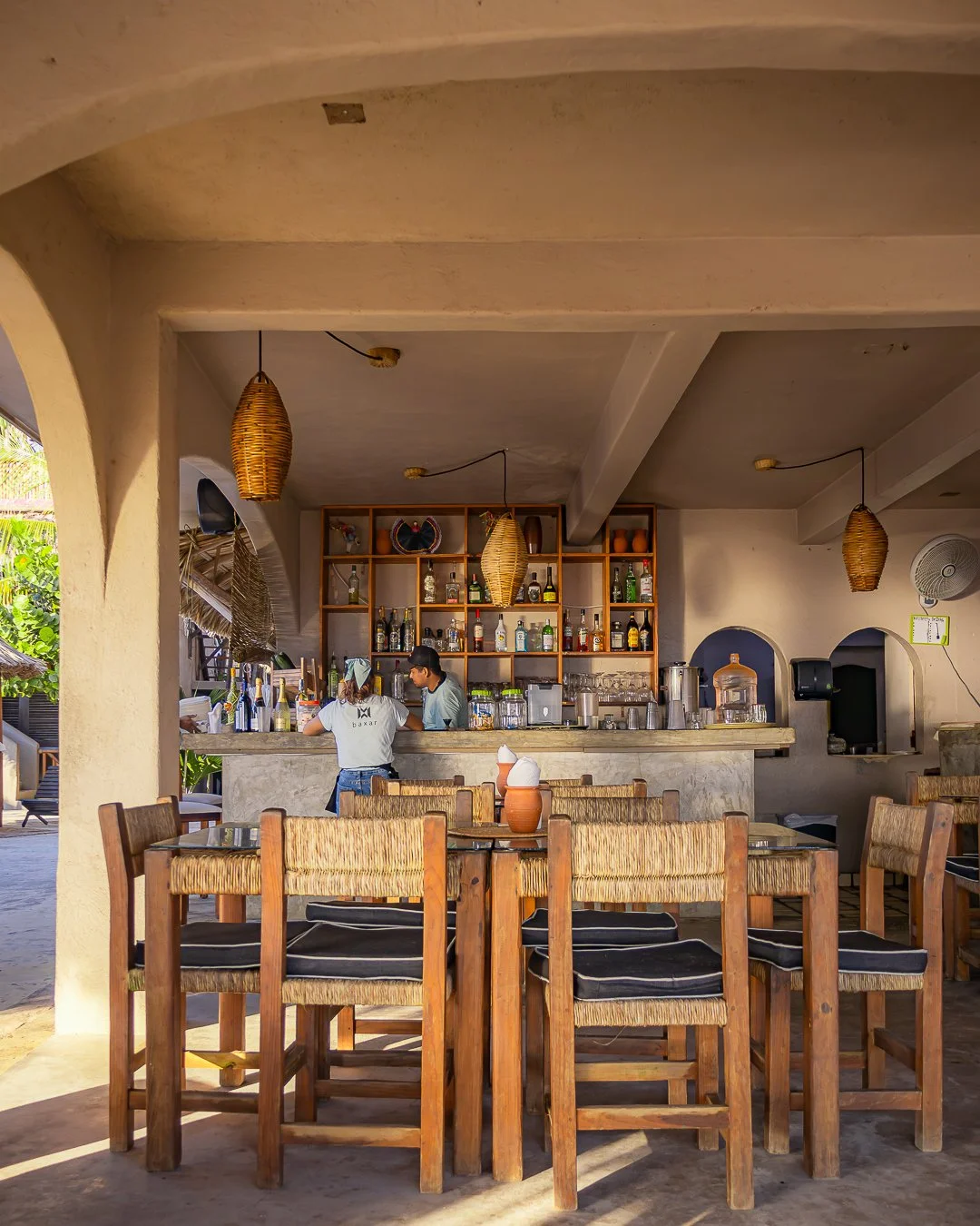 Xepina Restaurante Pie de la cuesta con mesas y sillas de madera, barra con estanterías llenas de botellas y utensilios, y dos empleados detrás de la barra, en un ambiente con decoración rústica y lámparas colgantes de mimbre.