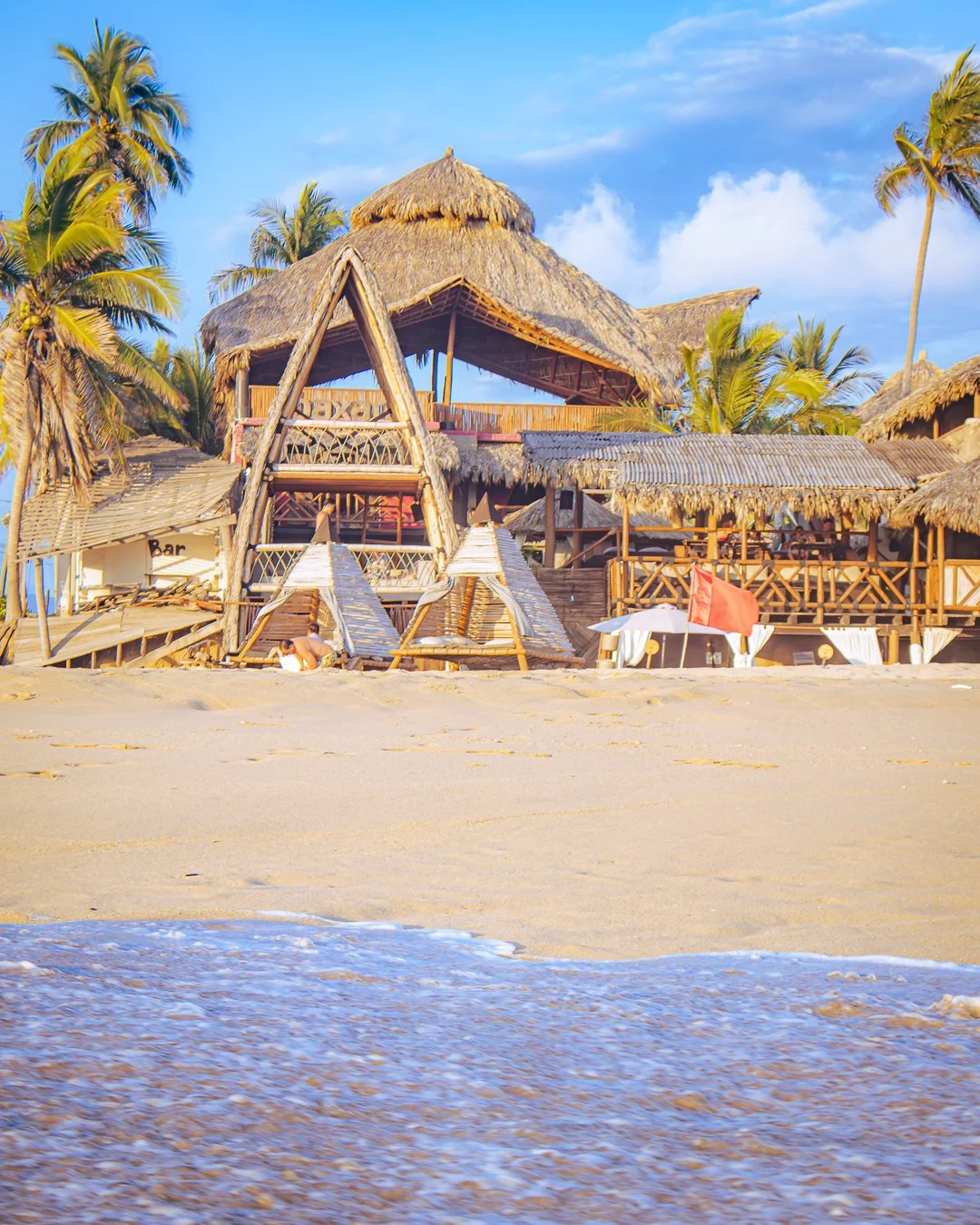 Casa en la playa con estructura de madera y techo de paja, rodeada de palmeras, con arena y mar al frente, en un día soleado y despejado. beach club del hotel baXar club de playa pie de la cuesta
