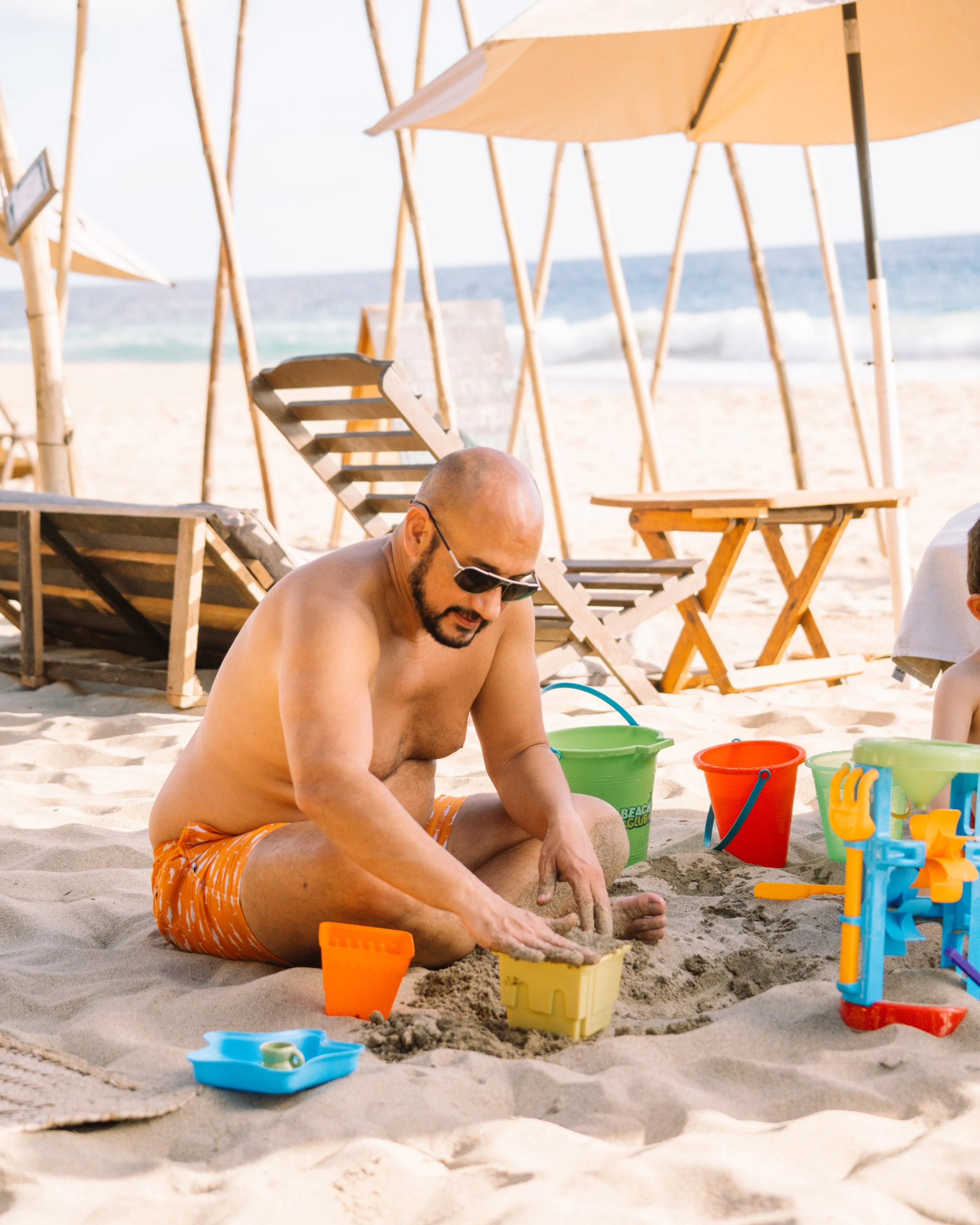 Un hombre sin camisa con gafas de sol y pantalones cortos anaranjados sentados en la arena jugando con arena; en el fondo se ven sombrillas, sillas y el mar en una playa. Hotel baXar pie de la cuesta