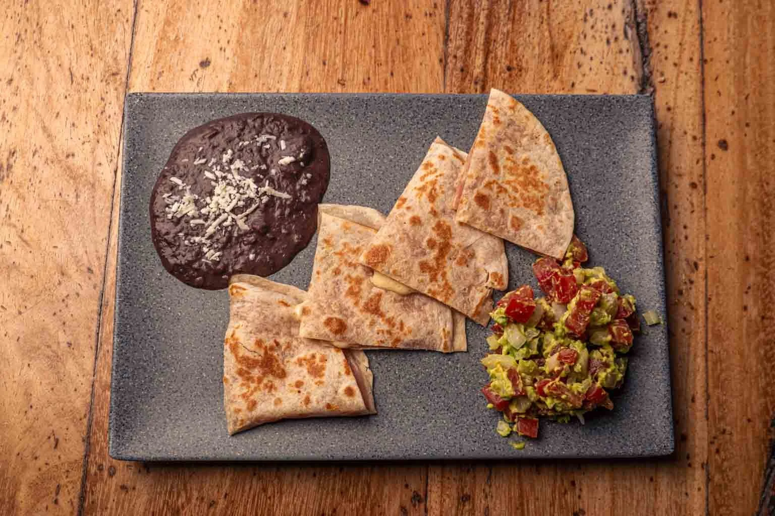Plato con cuatro quesadillas en un plato rectangular gris, acompañado de frijoles negros con queso en la esquina superior izquierda y pico de gallo en la esquina inferior derecha, sobre una mesa de madera.