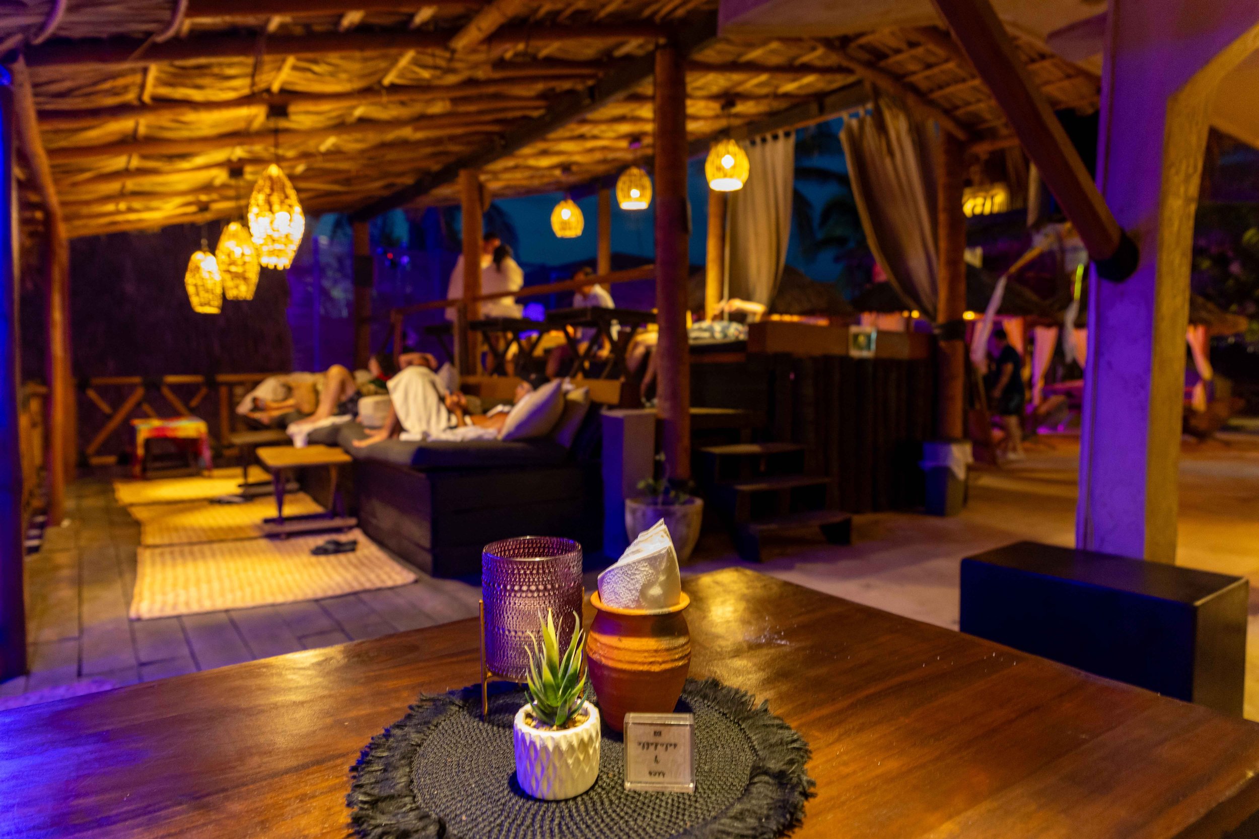 Interior de un restaurante con decoración rústica en madera, con mesas, sillas y lámparas de mimbre, ambiente cálido y exclusivo day pass club de playa pie de la cuesta. Hotel baXar