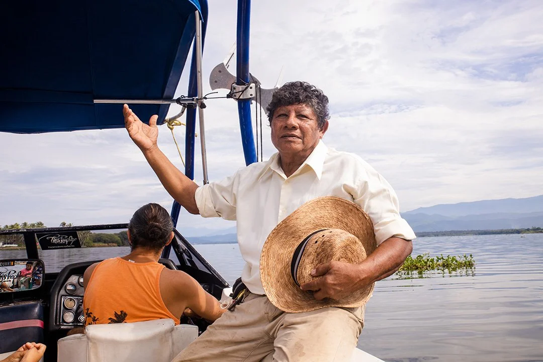 Hombre en barco en un río con un sombrero en la mano y una mujer conduciendo el barco, montañas laguna de coyuca, cielo nublado. Hotel boutique playa pie de la cuesta, baXar Acapulco
