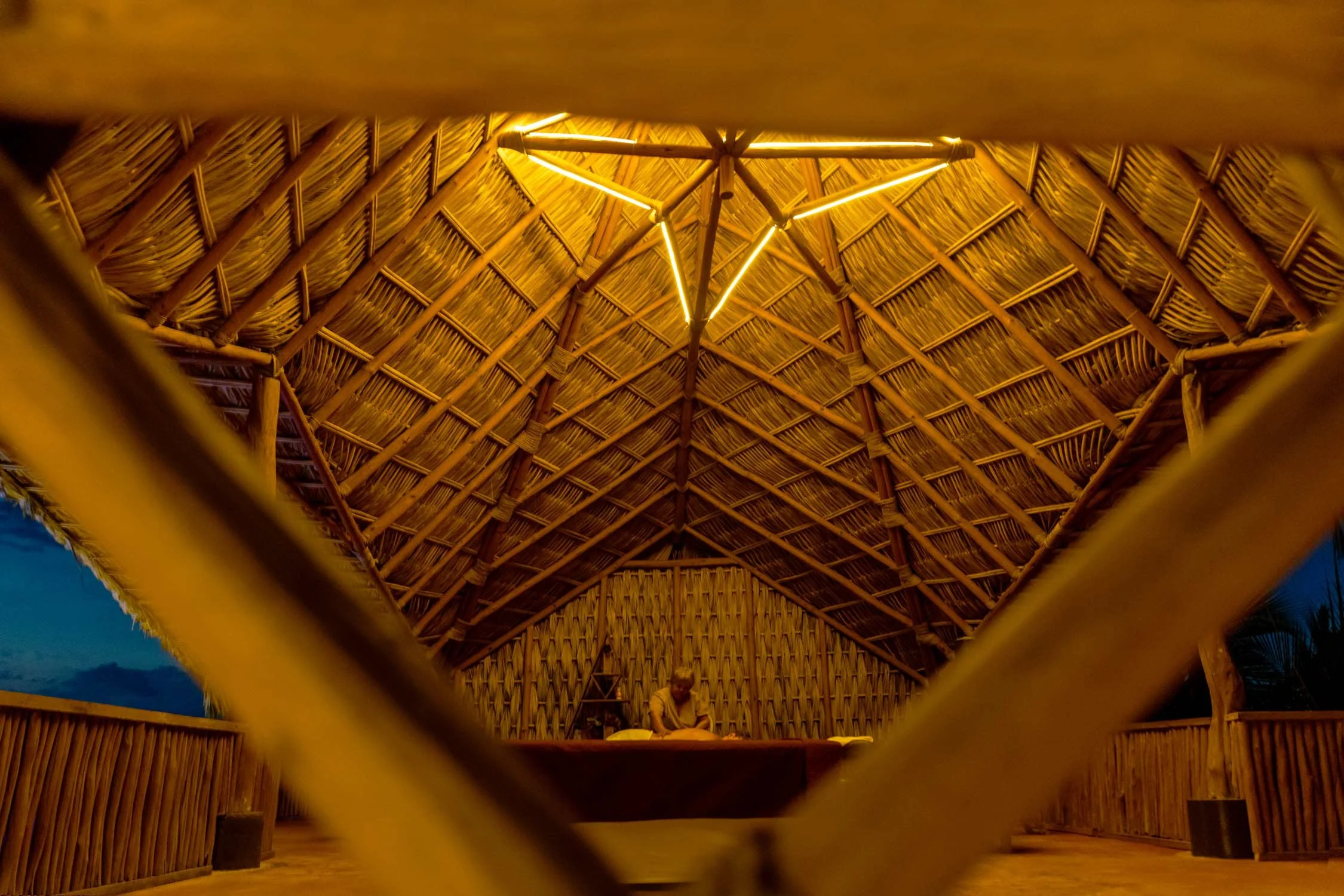 Vista del interior del spa palapa de baxar hotel wellness acapulco, palapa con brazos de madera, iluminada por luces cálidas, con una persona sentada en el fondo, probablemente en un lugar de descanso o reunión.