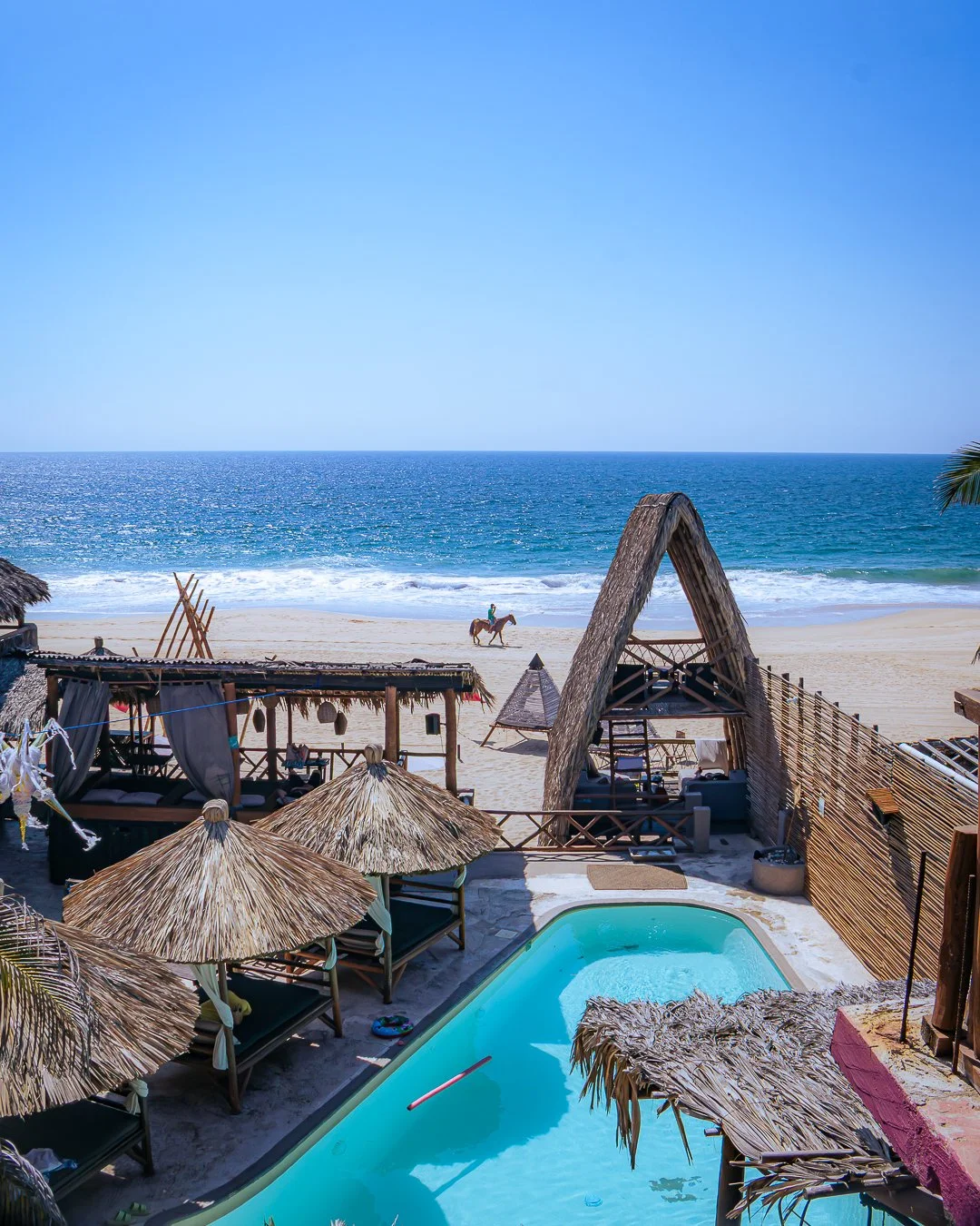 Vista de una Playa Pie de la Cuesta, mar azul y cielo despejado, con una cabaña de madera y sombrillas de paja en primer plano, y una piscina pequeña en el área de descanso. En la playa, un caballo con jinete pasea cerca del agua. Hotel baXar