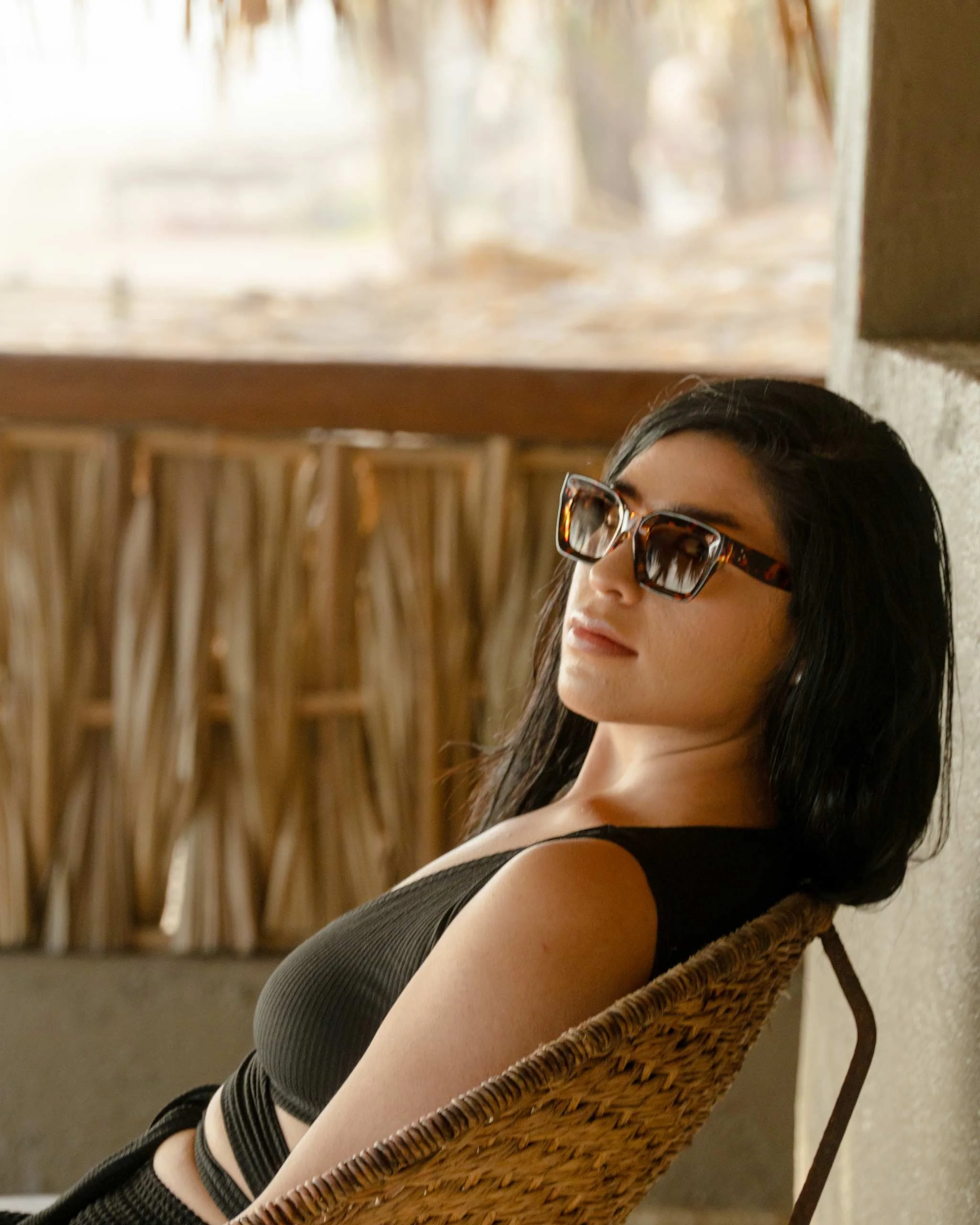 Mujer con gafas de sol de estilo tortoise, cabello largo y oscuro, vestida con un top negro, sentada en una silla de mimbre, en un ambiente con paredes de barro y cañas en el fondo. Hotel boutique baXar playa Pie de la cuesta Acapulco