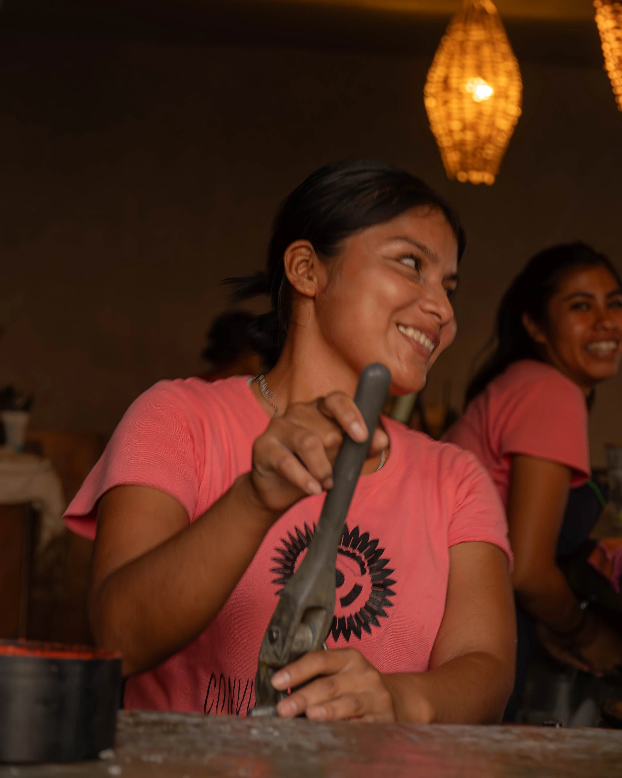 Mujer sonriendo, usando camiseta rosa de hotel baXar, sosteniendo un objeto en sus manos, en un ambiente cálido con lámparas colgantes de luz amarilla, detrás otra mujer también sonriente. club de playa day pass baXar