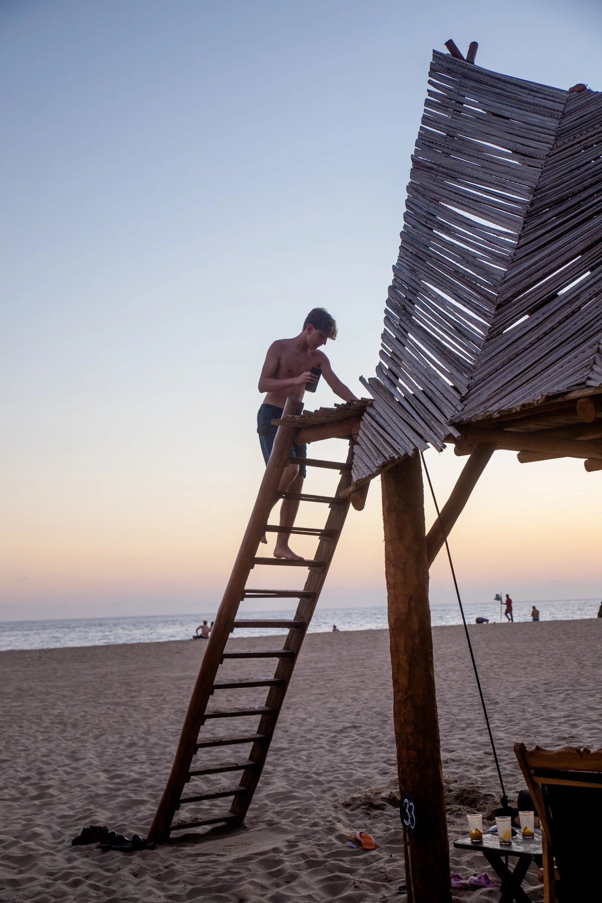Joven sin camisa en una estructura de madera en la playa al atardecer, con varias bebidas y sandalias en el suelo.  Hotel boutique baXar playa Pie de la cuesta Acapulco