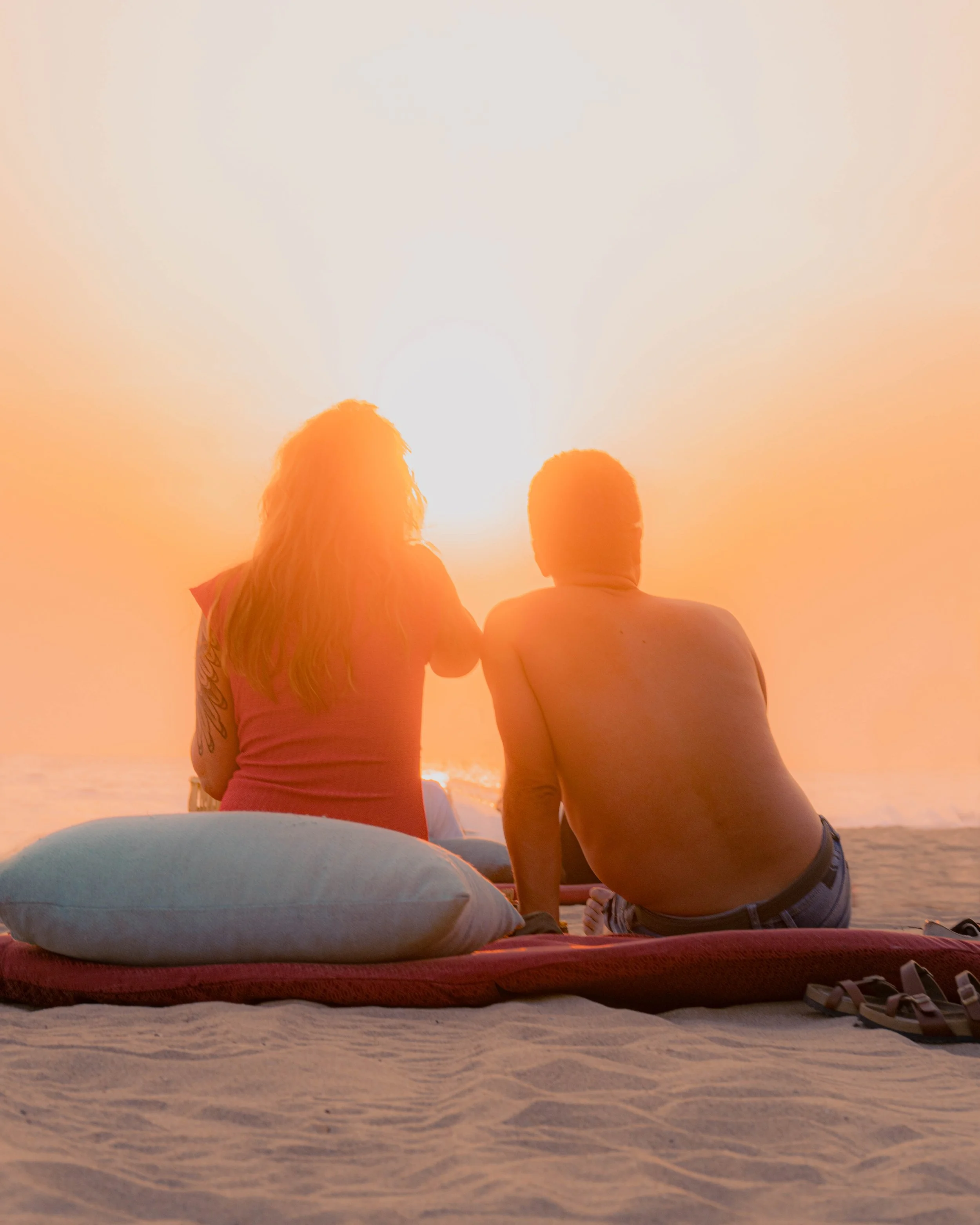 Dos personas de espaldas, un hombre y una mujer, sentados en playa pie de la cuesta durante la puesta del sol, con una almohada y sandalias en primer plano. baxar hotel acapulco