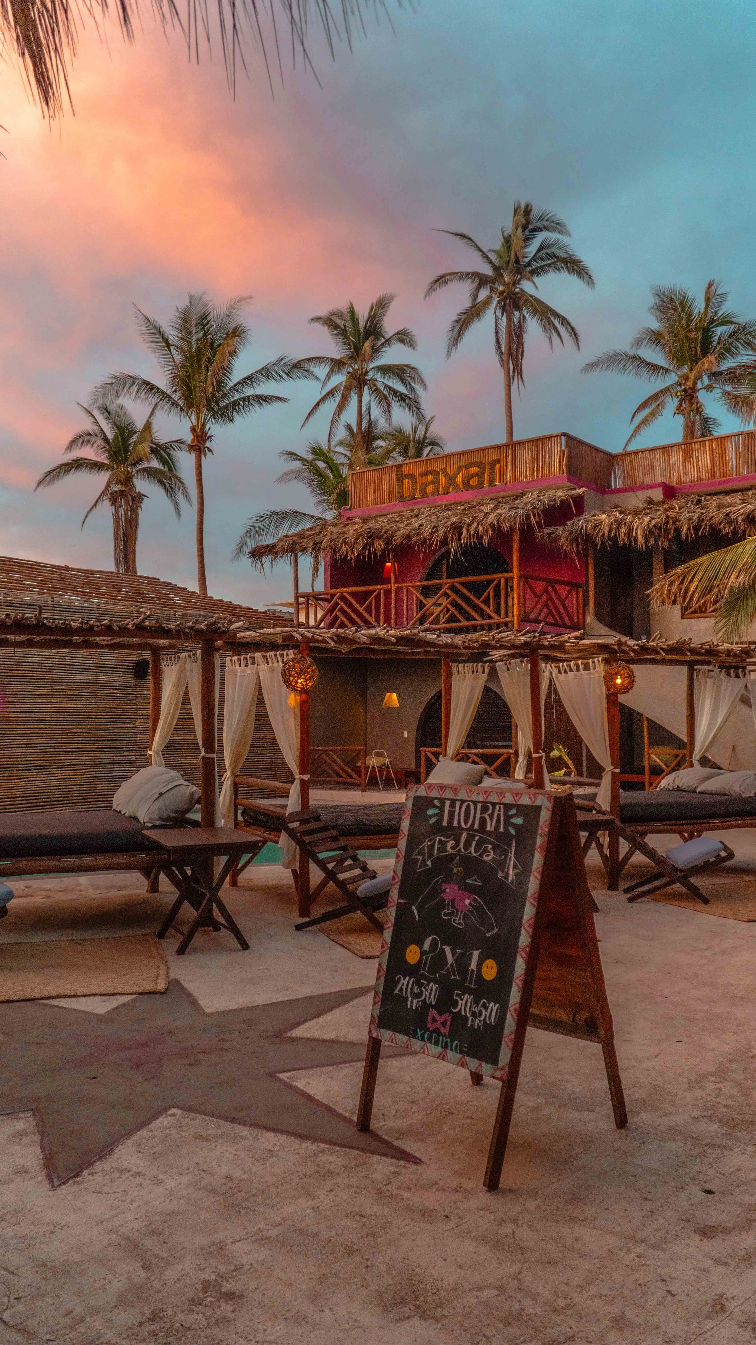 Escenario en una playa al atardecer con cabañas de madera, palmeras y una pizarra con mensaje en español que dice 'Hora Feliz, 2x1, 21:30 a 50/50 pm, en Xperience' hotel baxar pie de la cuesta 