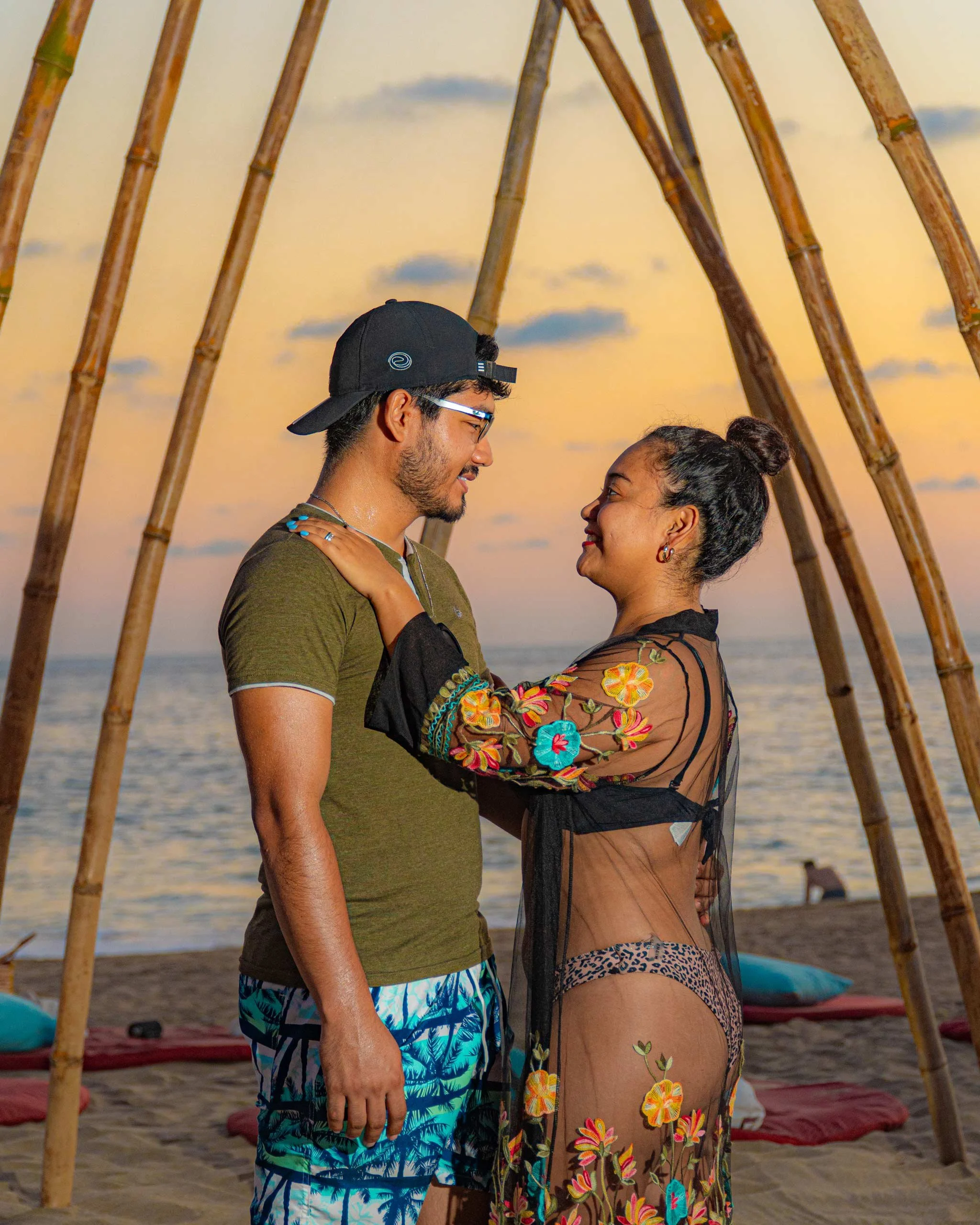 Una pareja feliz en una playa al atardecer, mirándose con amor, con un arco de madera de fondo y arena en el suelo.  Hotel boutique baXar playa Pie de la cuesta Acapulco