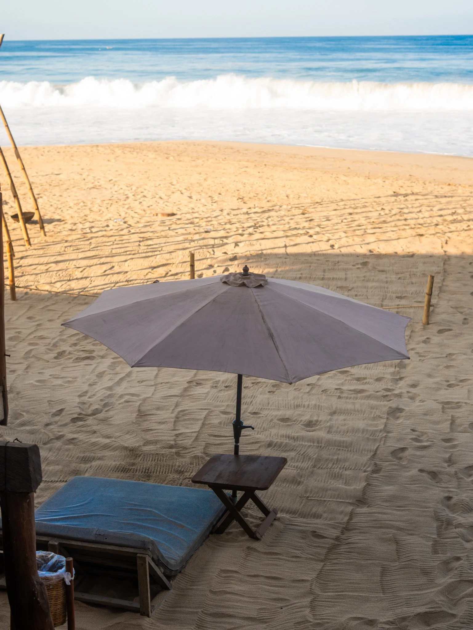 Vista de una playa con arena, una sombrilla gris en primer plano, y el mar con olas en el fondo.  Hotel boutique baXar club de playa Pie de la cuesta Acapulco. Beach club day pass