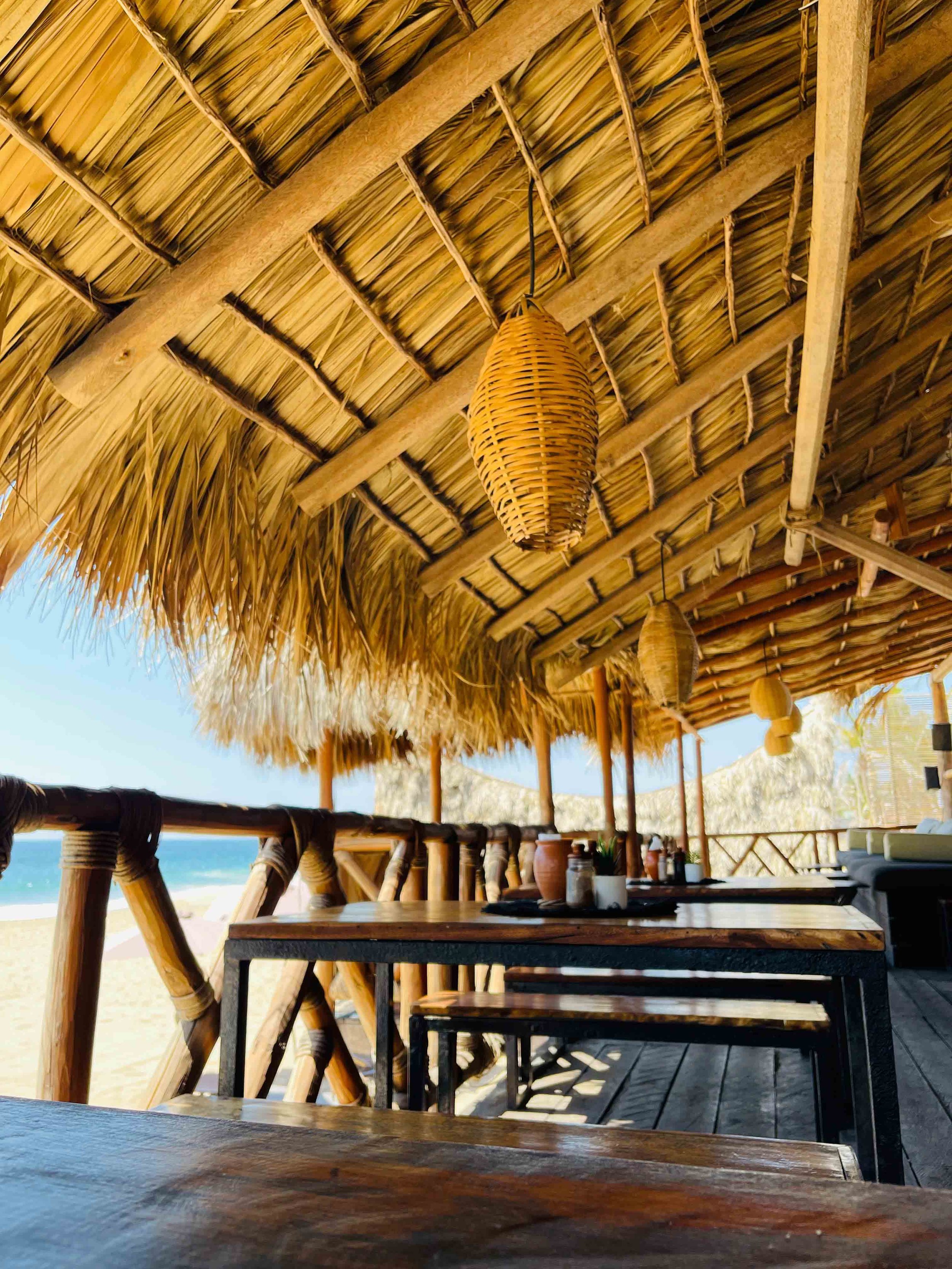 Terraza de restaurante con techadura de caña y lámparas de papel, vista a la playa con el mar y arena al fondo, mesas de madera con utensilios sobre ellas. hotel baxar pie de la cuesta 