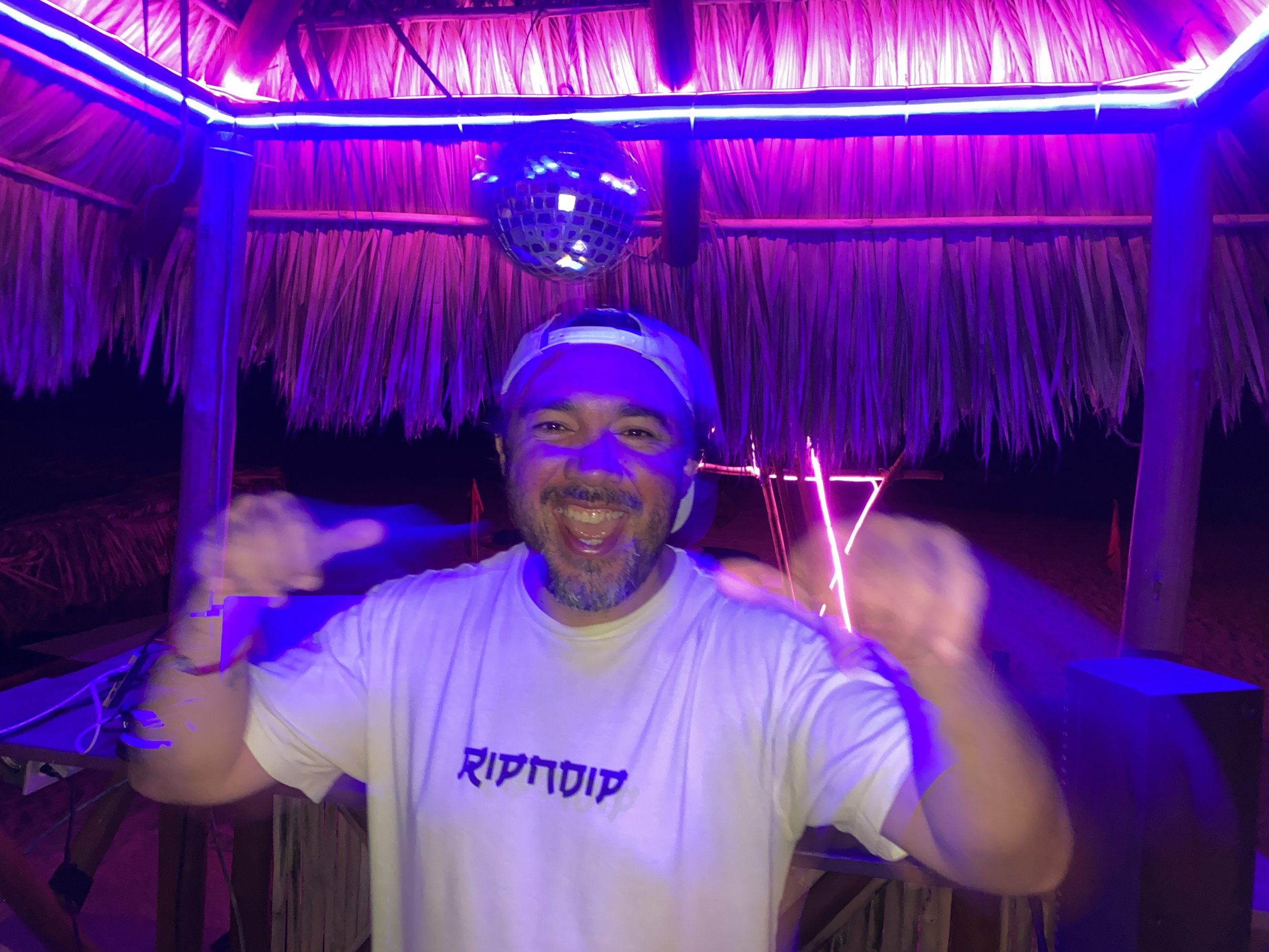 DJ Cool sonriendo y vestido con camiseta blanca en un lugar con iluminación de neón púrpura y fondo de palapa beach acapulco. - baXar Pie de la Cuesta