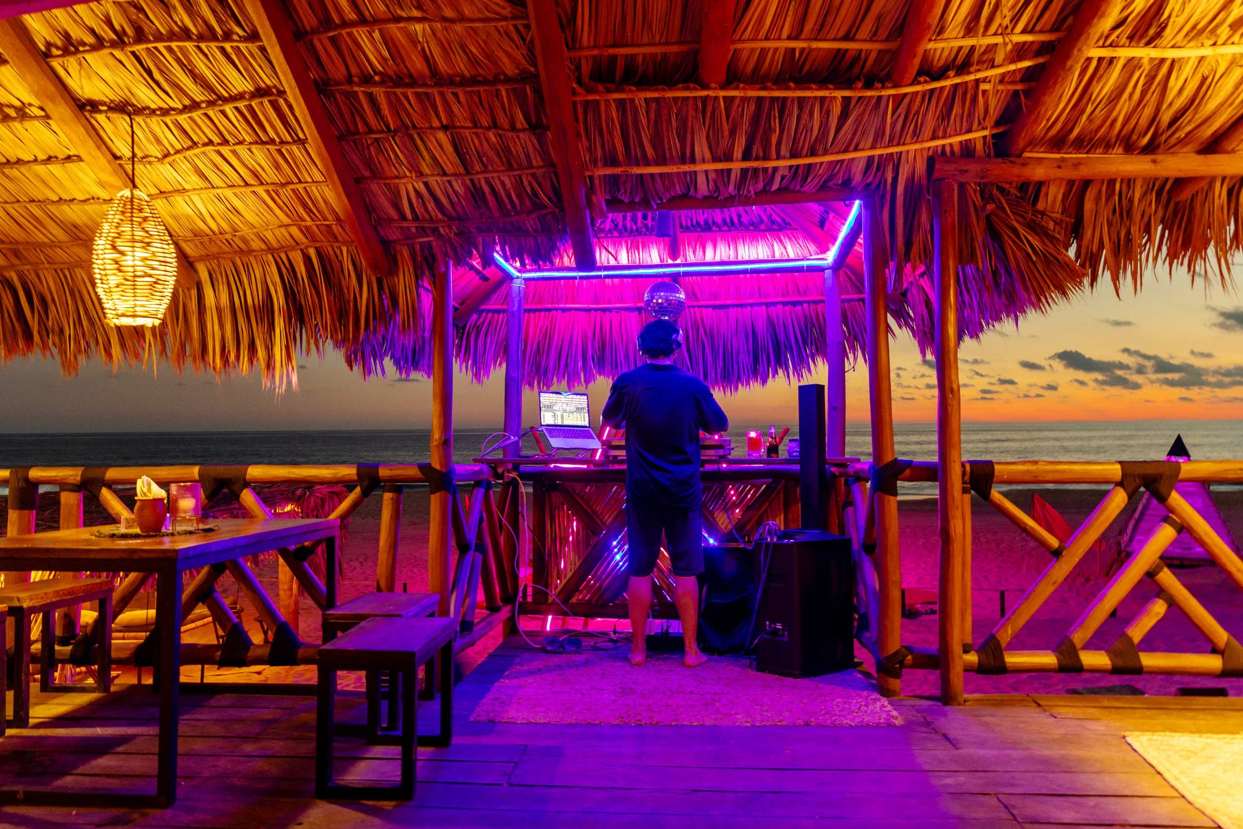 Atardecer en un playa pie de la cuesta, bar de palapa con luces de neón moradas y un DJ tocando música, hotel boutique baXar Acapulco