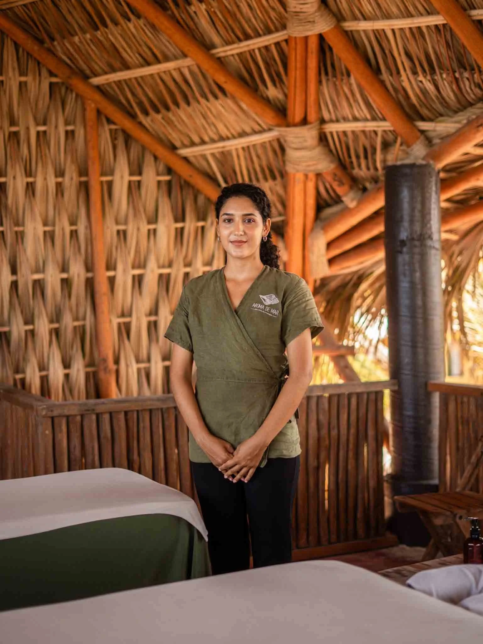 Mujer de cabello rizado y piel morena, sonriendo, en uniforme verde, en una estructura de madera y paredes de caña, probablemente en un espacio de relajación o spa. hotel baxar pie de la cuesta 