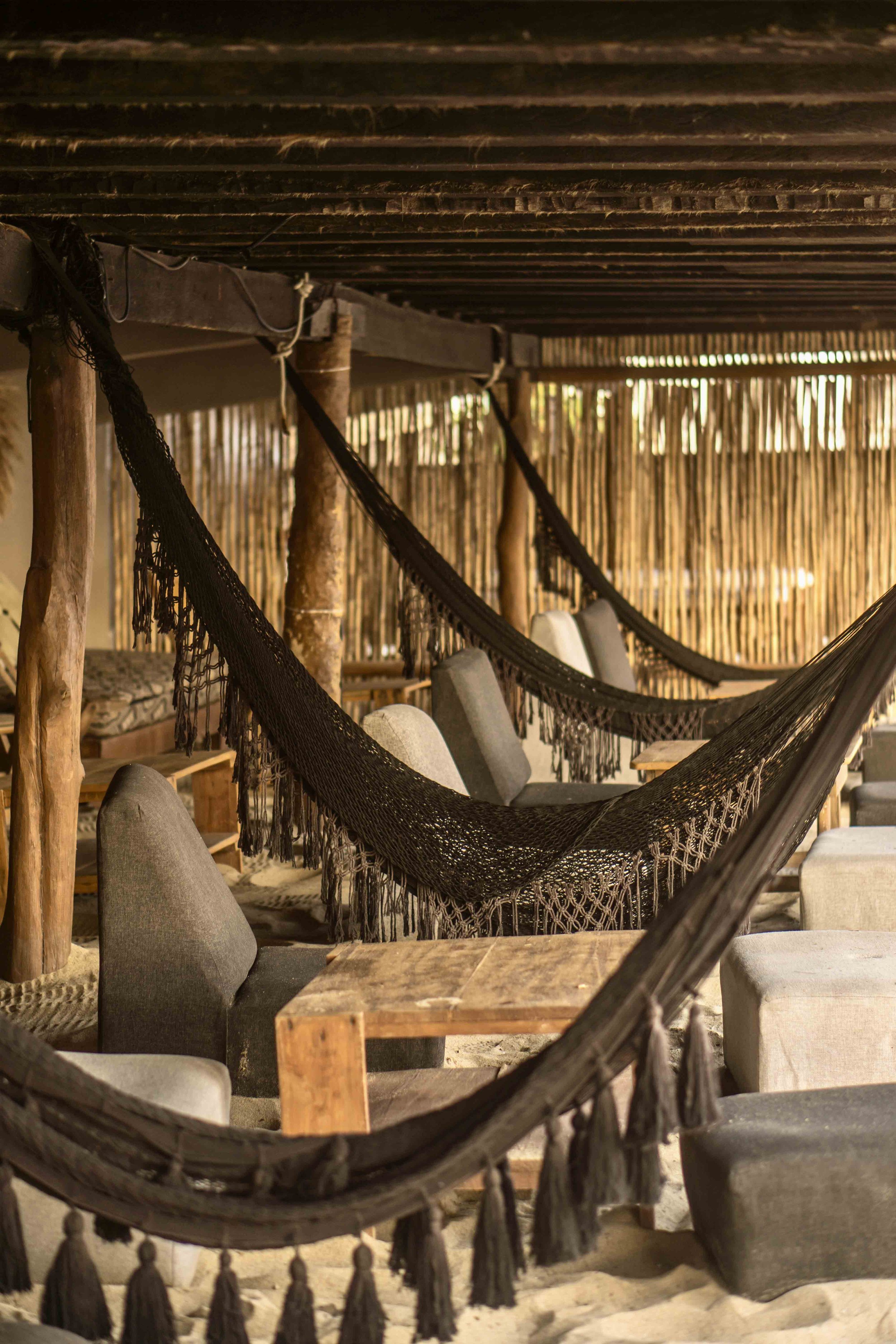 Cabaña de playa con hamacas de malla negra y asientos con cojines en la arena, paredes de madera y techo de láminas.