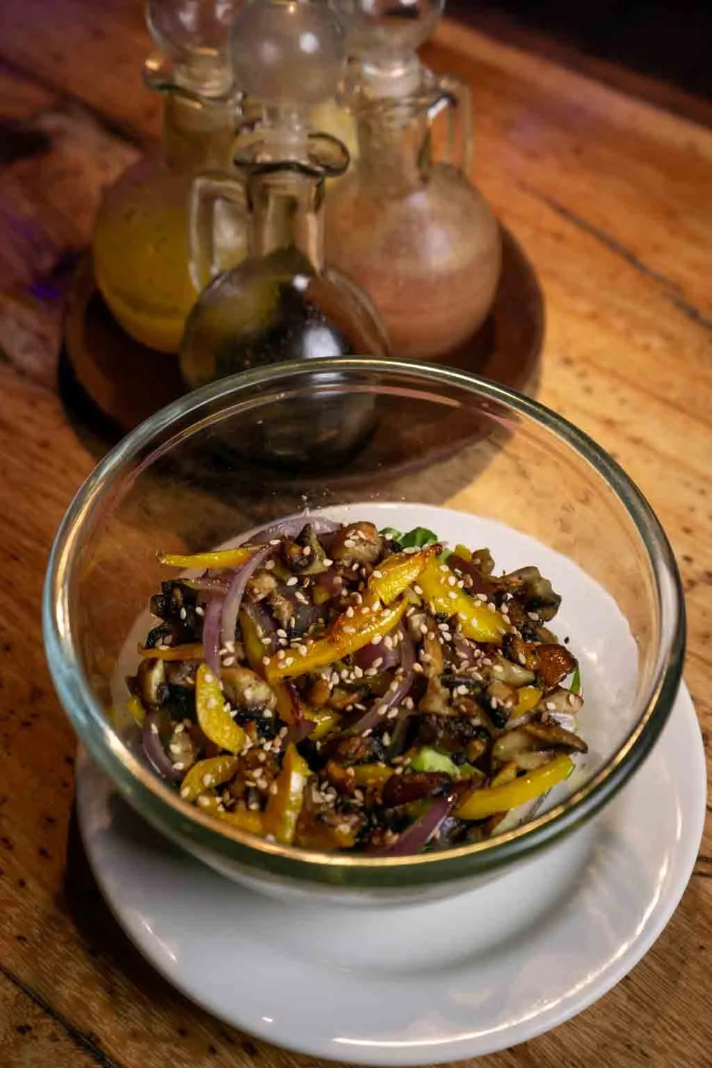 Ensalada en un vaso de cristal con semillas y vegetales, en una mesa de madera con pequeños frascos de cerámica y vidrio en el fondo.