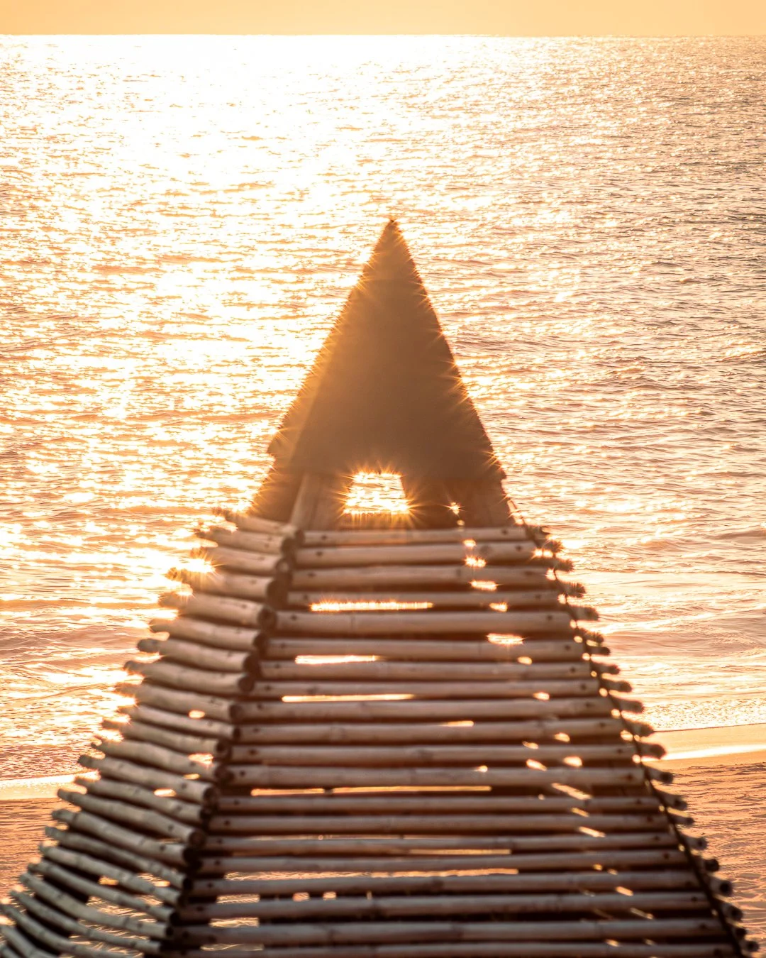 Estructura de madera en forma de triángulo sobre la playa, con el sol brillante de fondo reflejándose en el mar. hotel boutique playa pie de la cuesta, baXar Acapulco