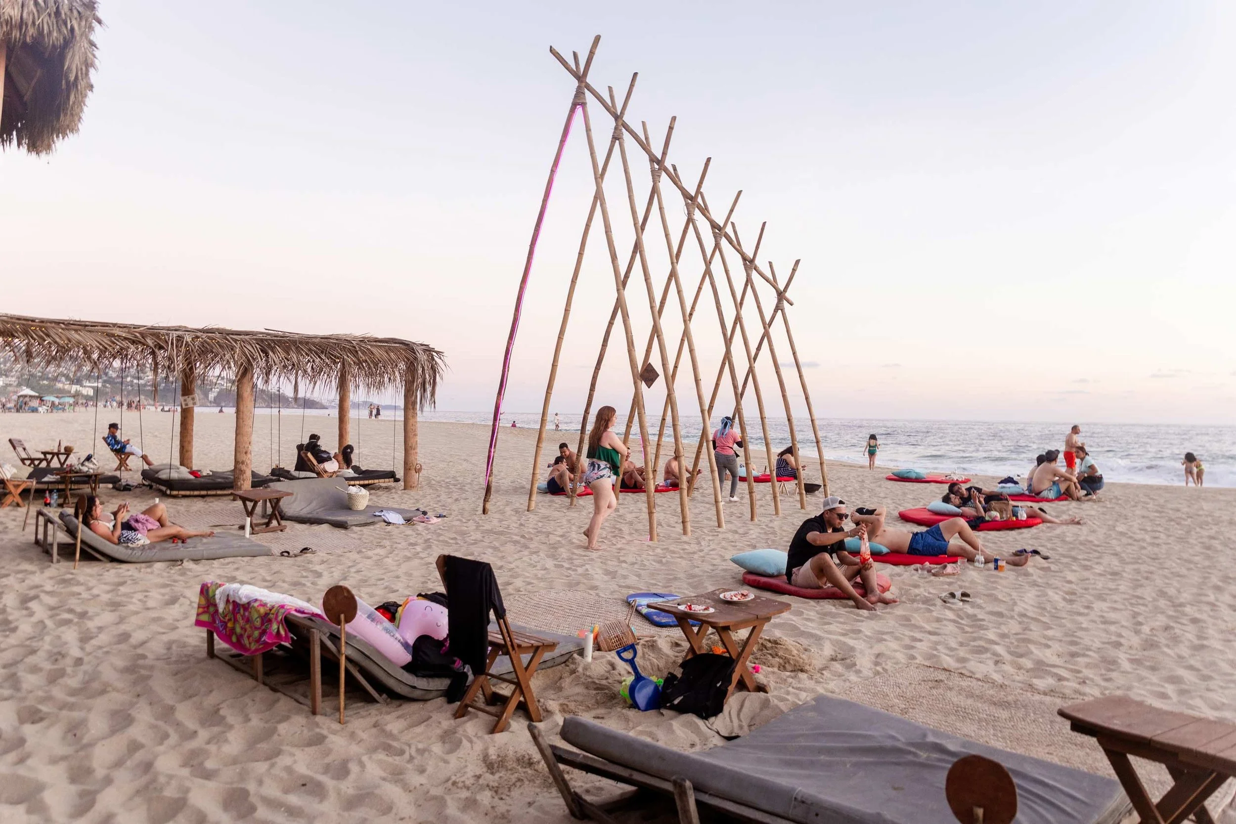 Playa con sombrillas de paja, personas en sillones y en la arena, estructuras de bambú, vista al mar y cielo despejado, ambiente tranquilo y relajado en la playa. day pass beach club de playa pie de la cuesta, hotel boutique baXar acapulco