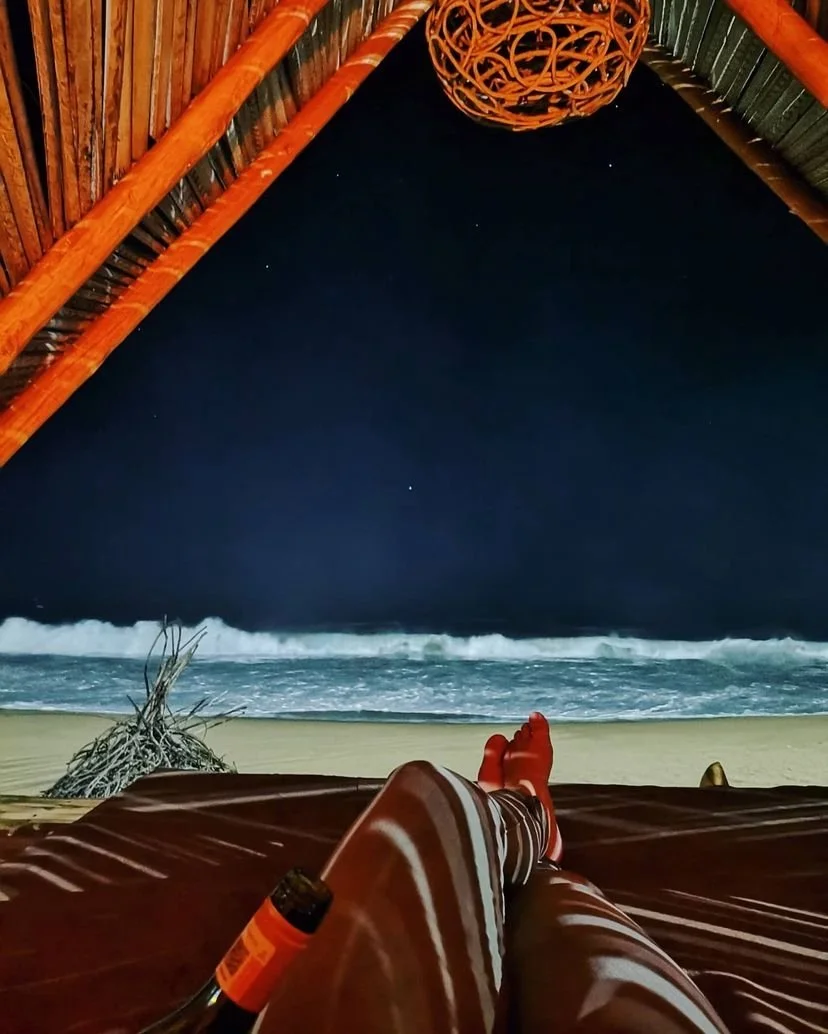 Vista desde una  Hotel boutique baXar playa Pie de la cuesta Ac de playa en la noche, con una persona descansando en una hamaca, foco en sus piernas y pies, con una botella en la mano, mirando hacia el mar con olas en la orilla y un cielo estrellado.