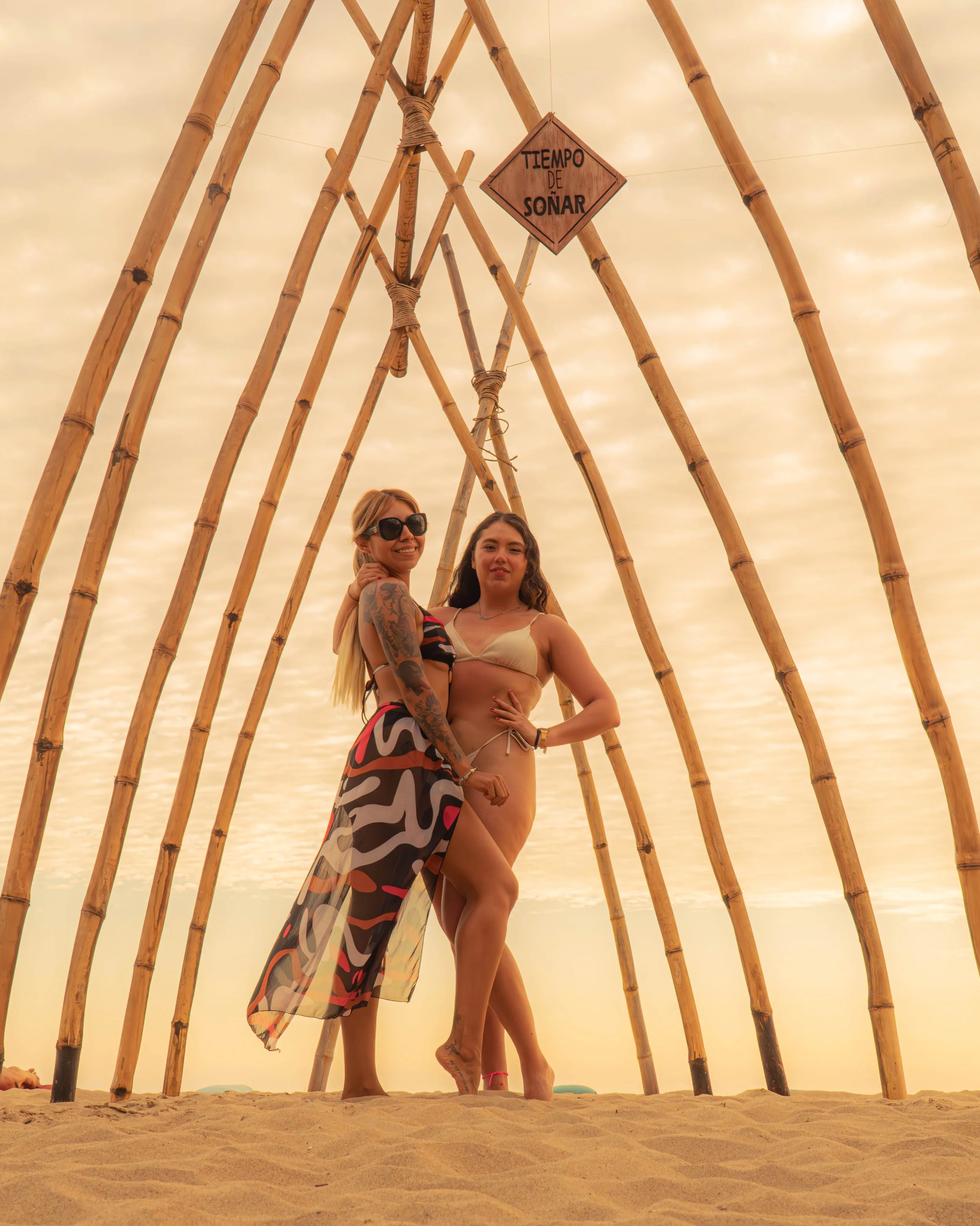 Dos mujeres de pie, una con gafas de sol y la otra sin, posando frente a un marco de bambú en la playa durante el atardecer, con un cartel que dice 'Tiempo de soñar' colgado encima. Hotel baXar playa pie de la cuesta