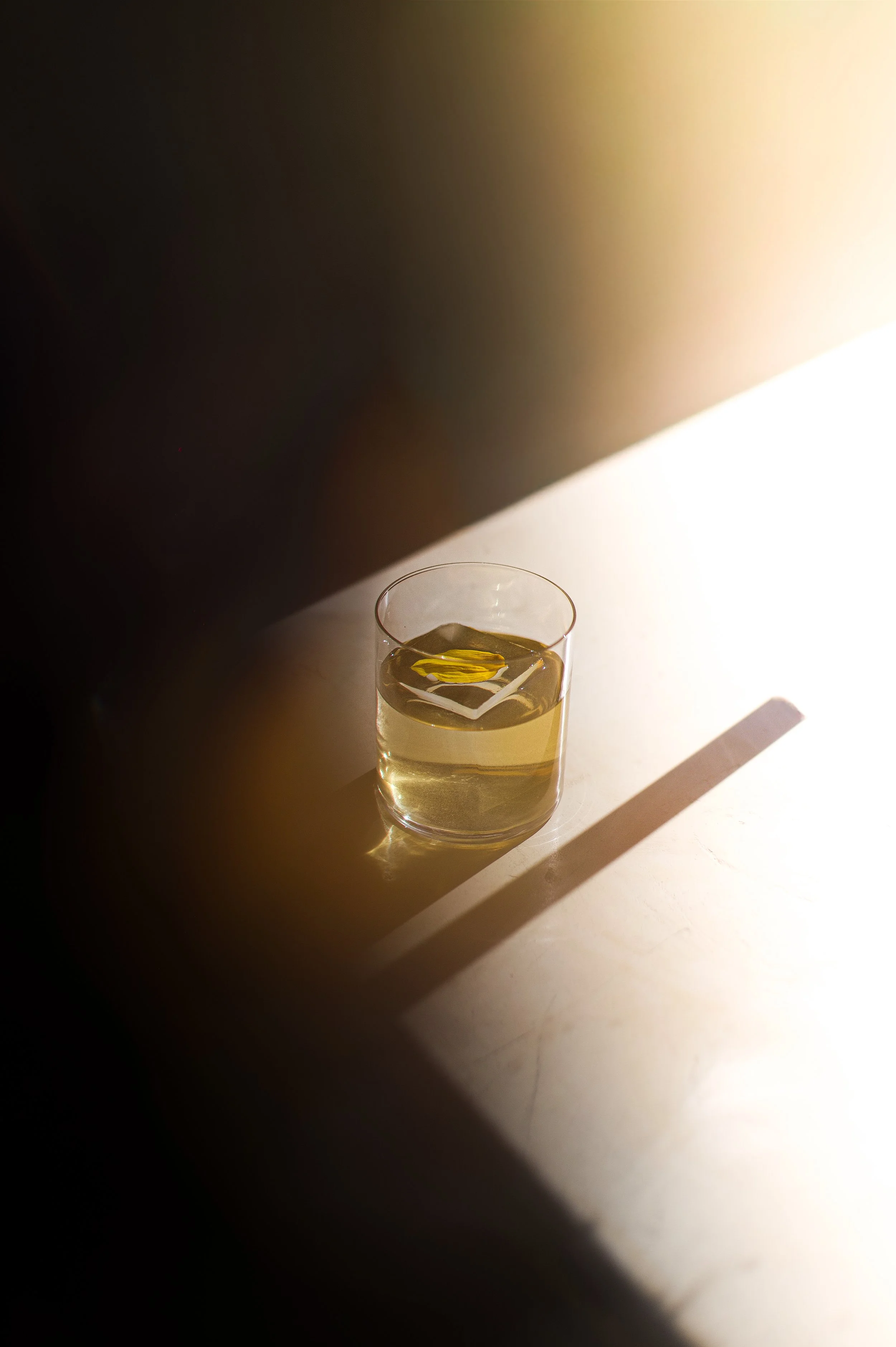 cocktail avec un cube de glace et quelques gouttes d'huile, sur une surface claire avec une forte source de lumière venant de la droite, créant une ombre longue.