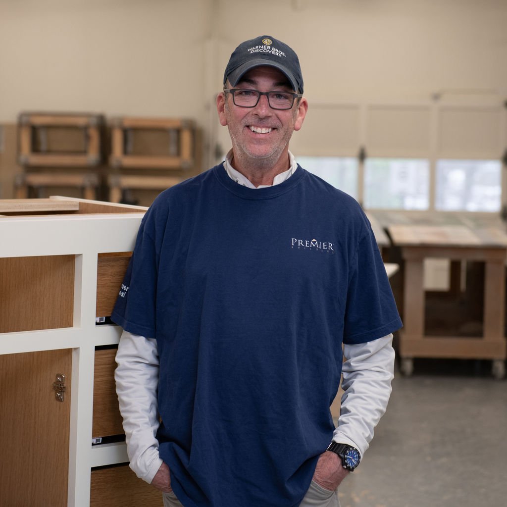 A middle-aged man with a beard, wearing a grey cap and a zipped navy blue jacket, sitting at a table with his arms resting on it, against a plain white background.