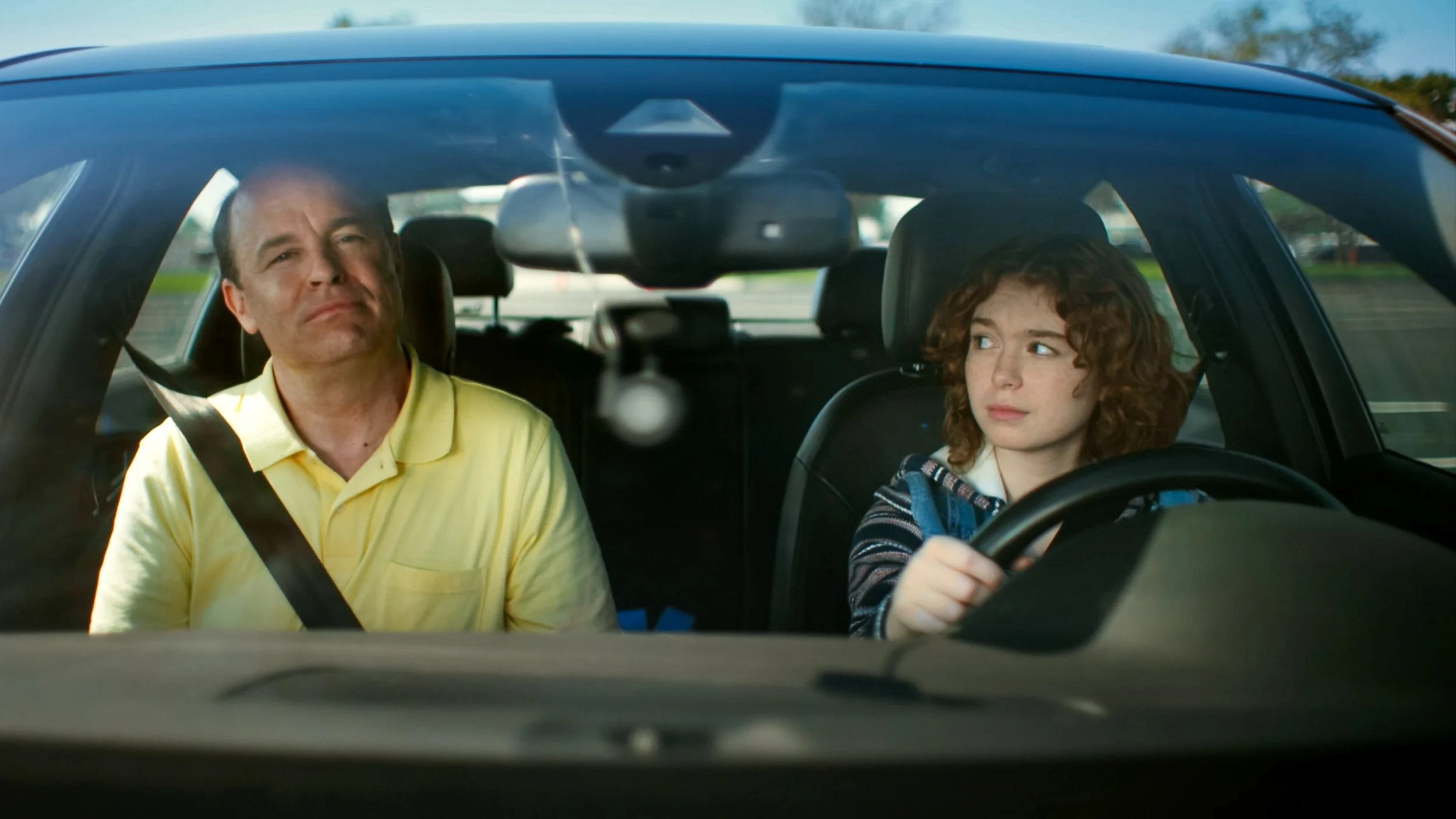 A man and a girl sitting inside a car, with the girl driving. The man is wearing a yellow shirt and the girl has curly red hair and is holding the steering wheel.