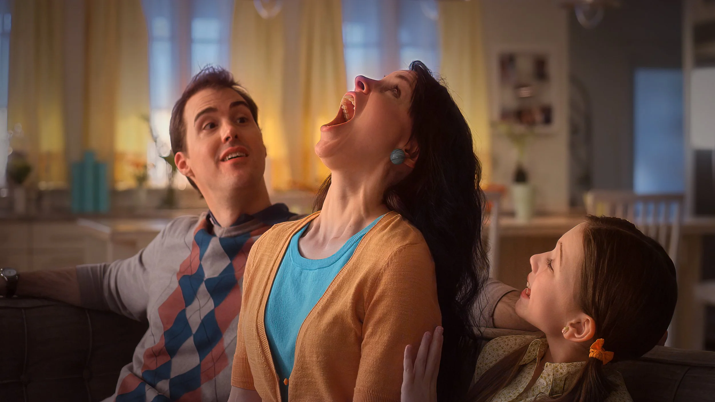 A family of three sitting on a couch with a brightly lit kitchen in the background. The woman in the center, with long dark hair and earrings, is opening her mouth wide in a playful or exaggerated expression. The young girl to her right, with pigtails and a bow, is smiling and looking up at her. The man on the left, with short dark hair, is looking at the woman with a surprised or amused expression.