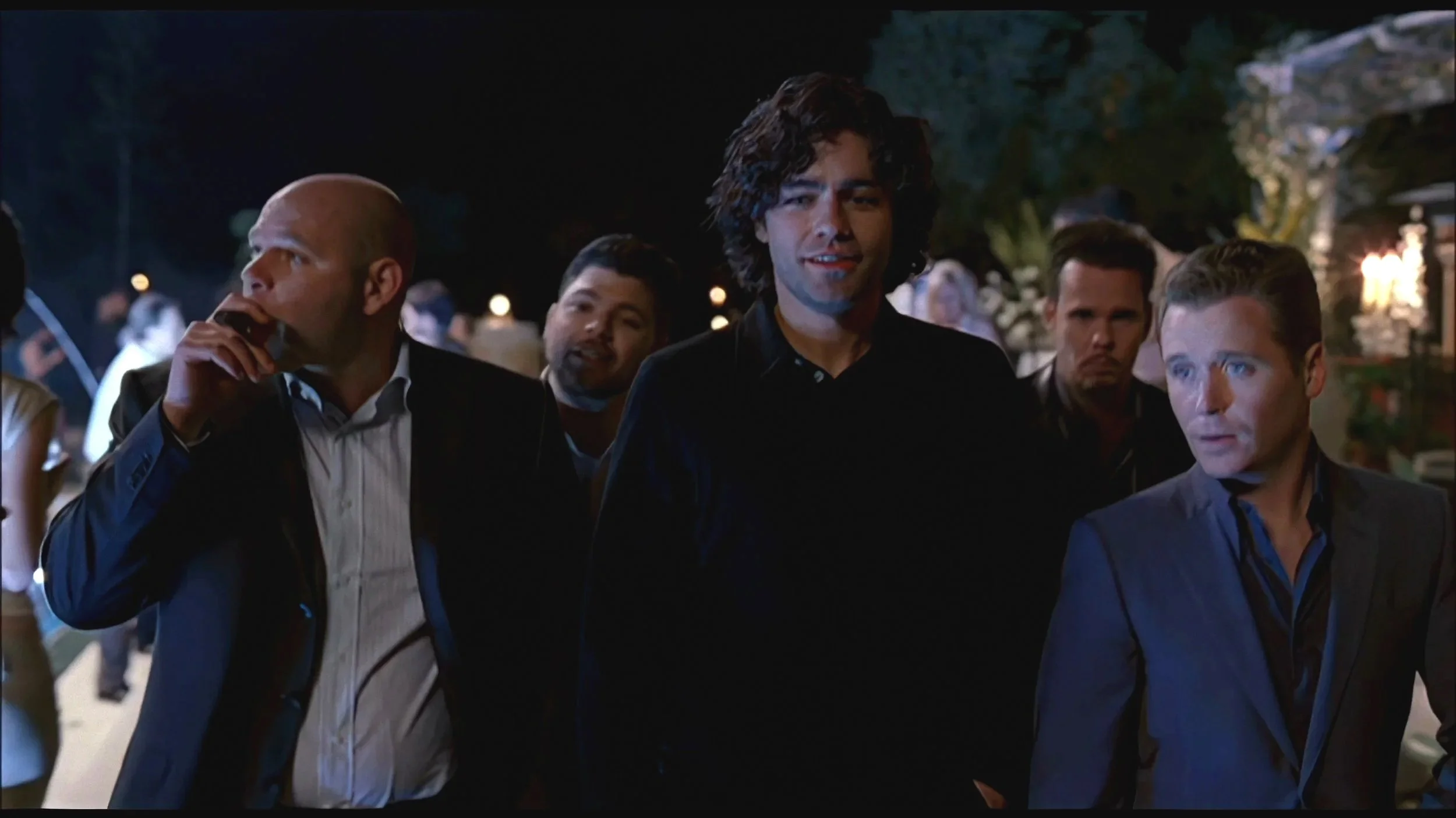 A group of men walking outdoors at night, dressed in formal attire, with some in suits, with blurred background lighting and trees.