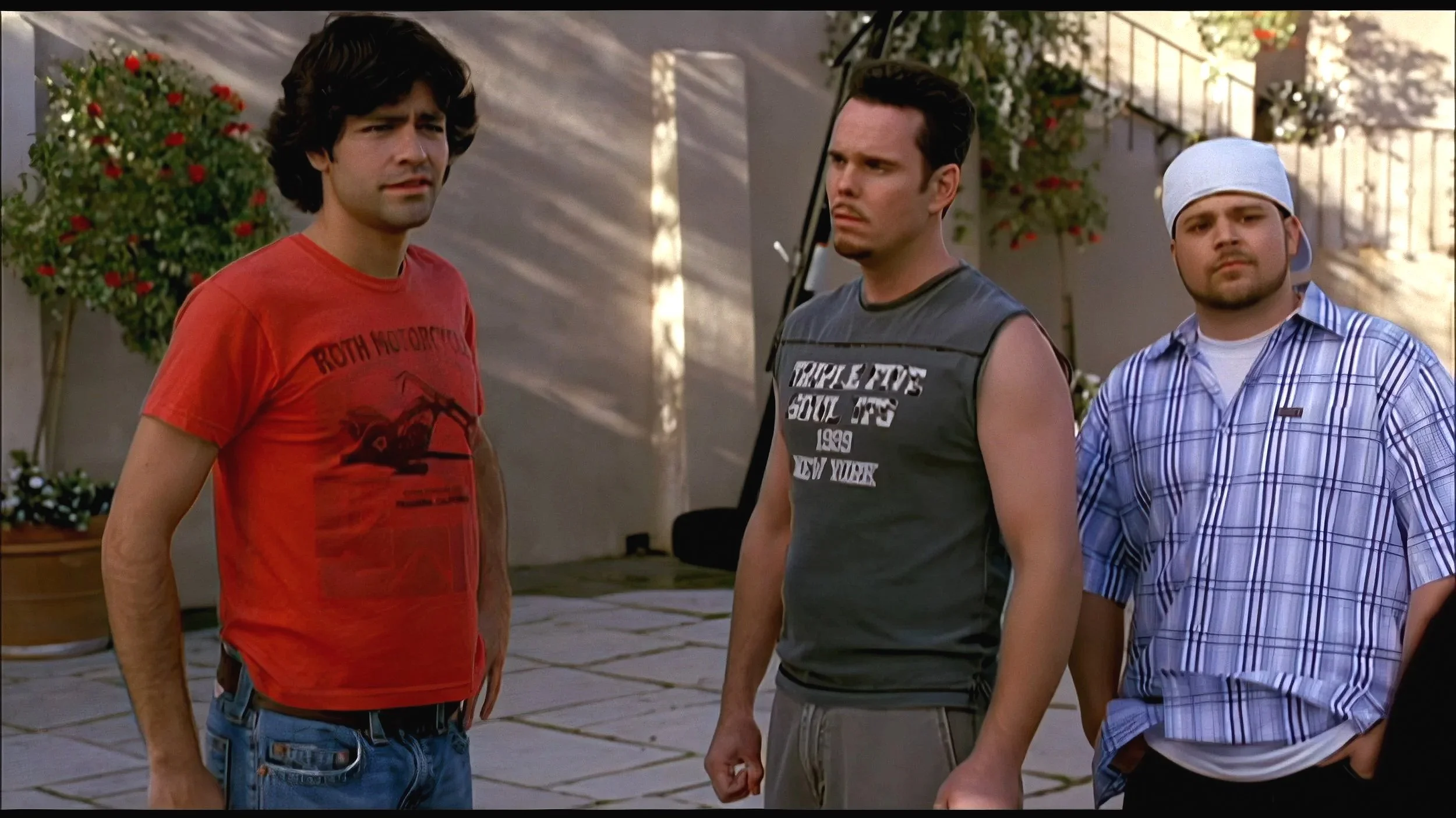 Three young men standing outdoors in casual clothing, with potted flowers and a staircase in the background.