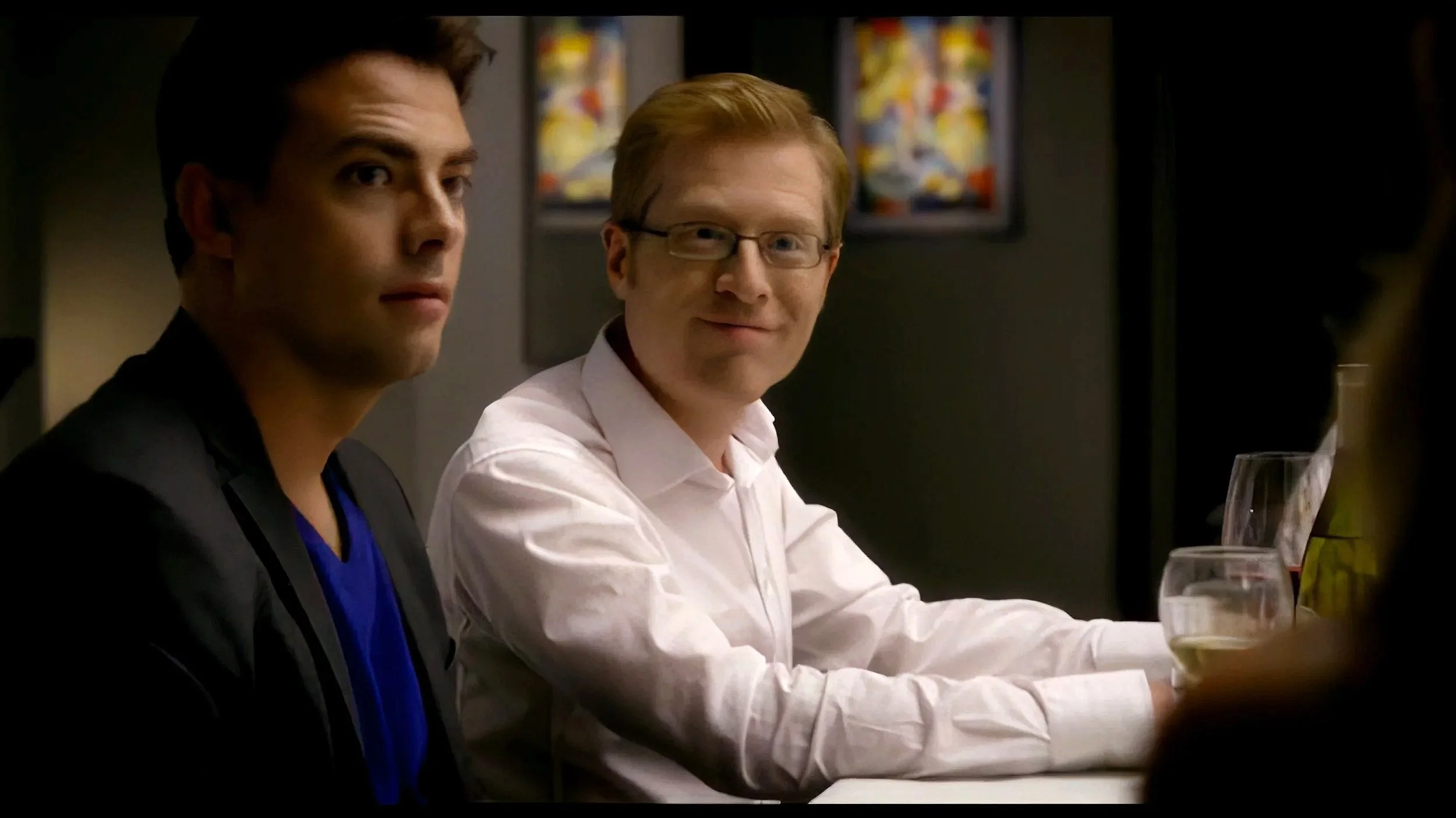 Two men sitting at a table, one with dark hair and wearing dark clothing, the other with red hair, glasses, and a white shirt, in a dimly lit setting, possibly a restaurant or bar, with drinks on the table.